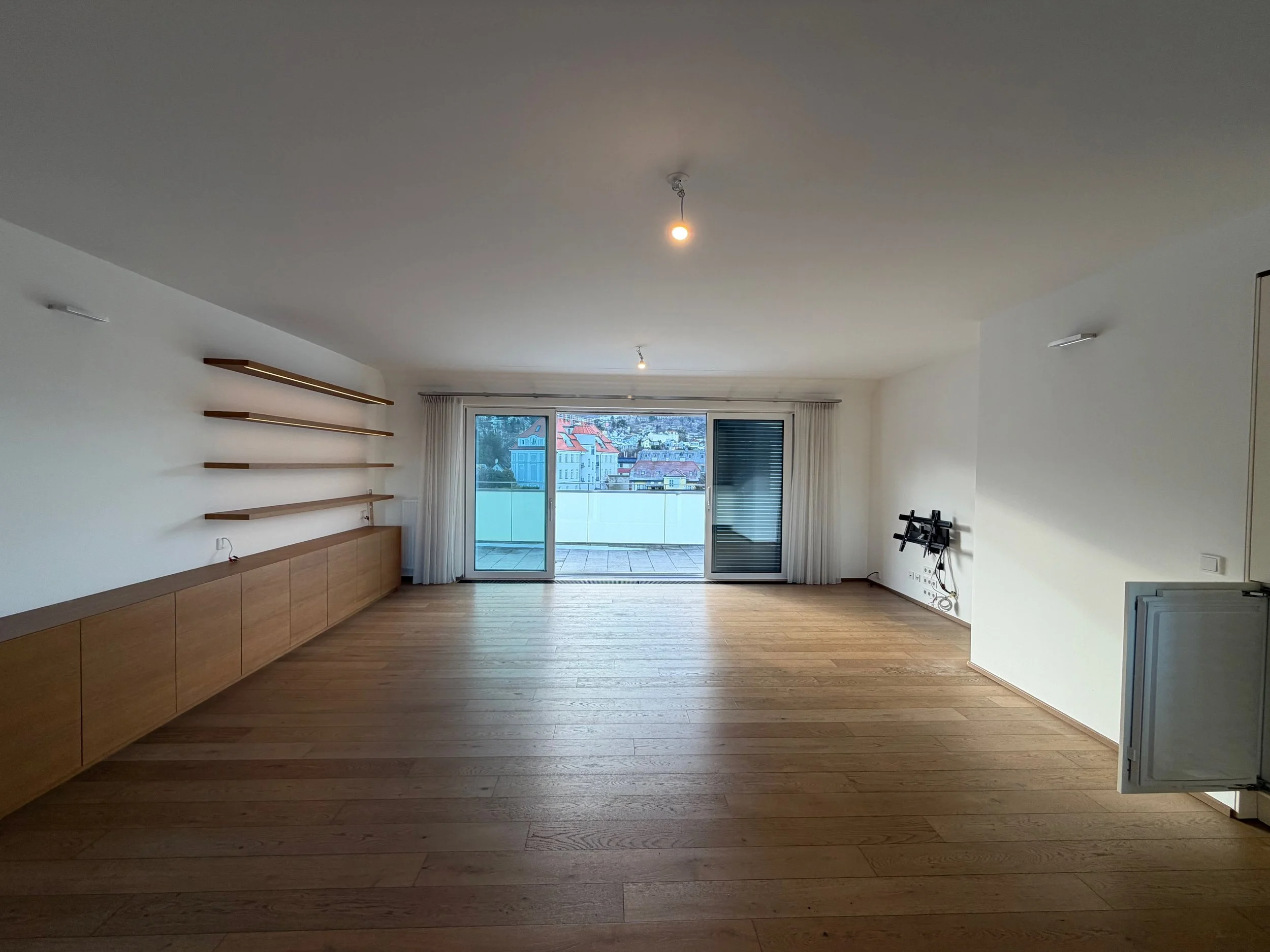 Leeres Wohnzimmer mit Holzboden, weißen Wänden, großen Fenstern mit Vorhängen, einigen Regalen an der Wand und einer kleinen schwarzen TV-Halterung auf der rechten Seite.