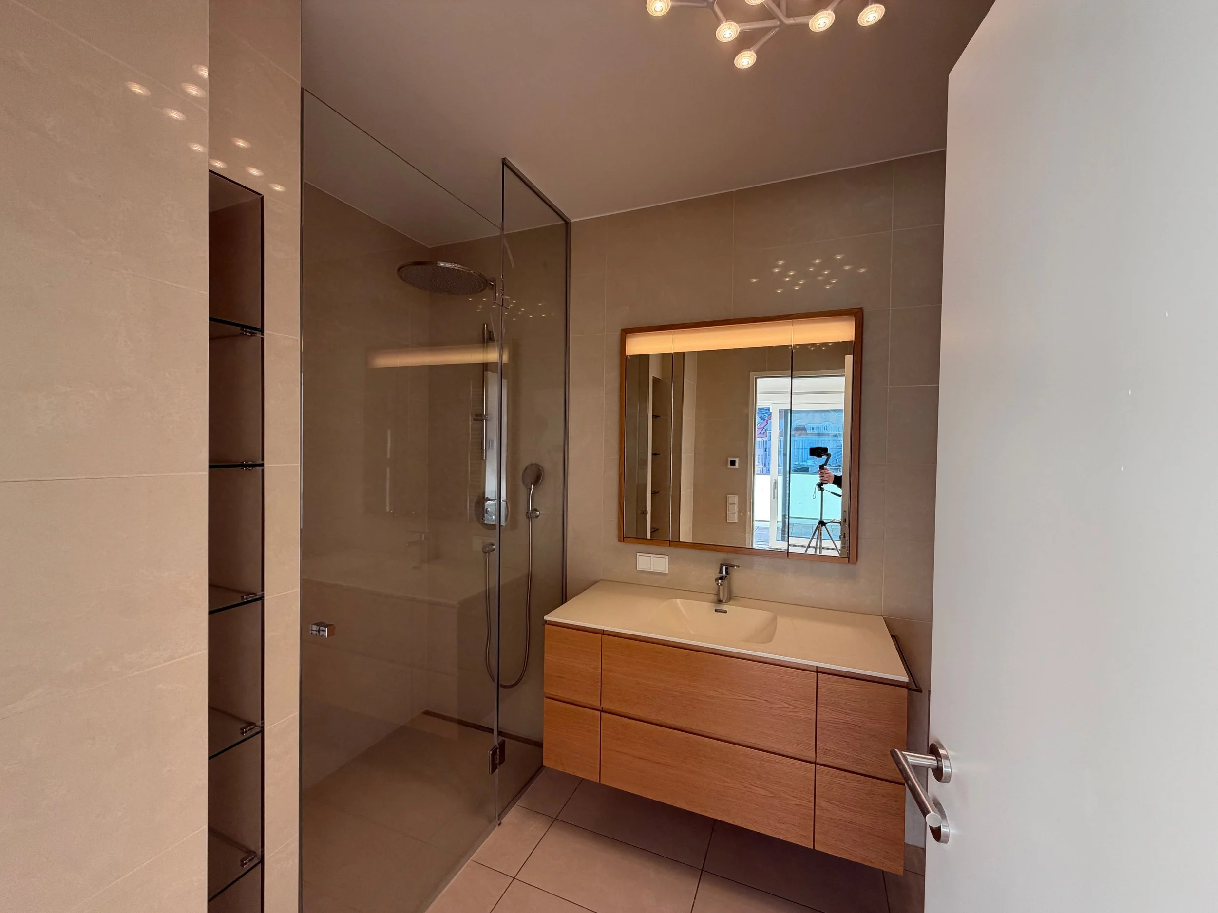 Moderner Badezimmerbereich mit beigem Wandfliesen, einem Waschtisch mit Holzschubladen, einem großen Spiegel und einer Duschkabine aus Glas mit Regendusche.