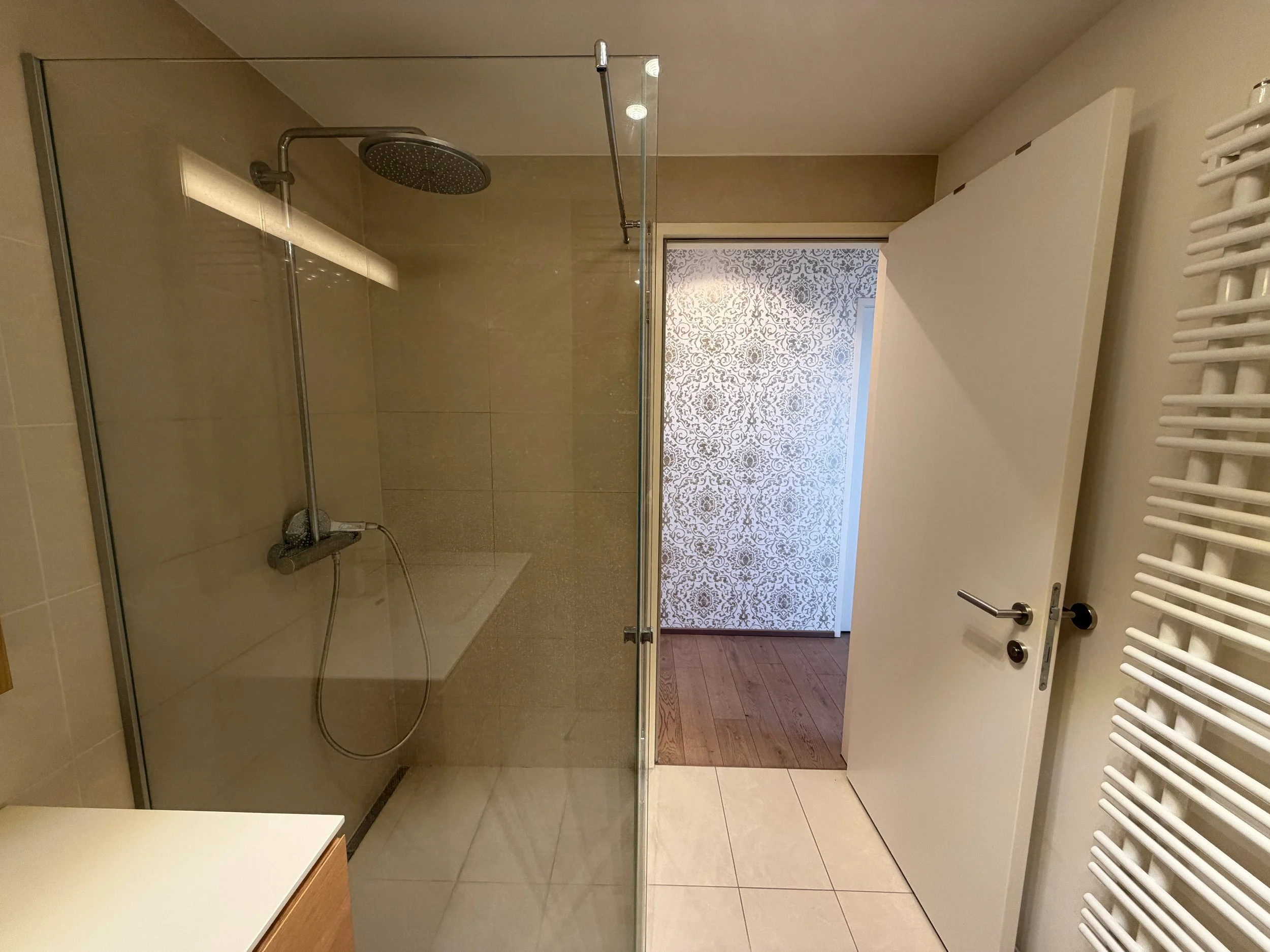 Badezimmer mit offener Duschkabine, Wandheizung und Holzboden im angrenzenden Raum, weiße Tür, beige Fliesen, dekorative Tapete im Flur.