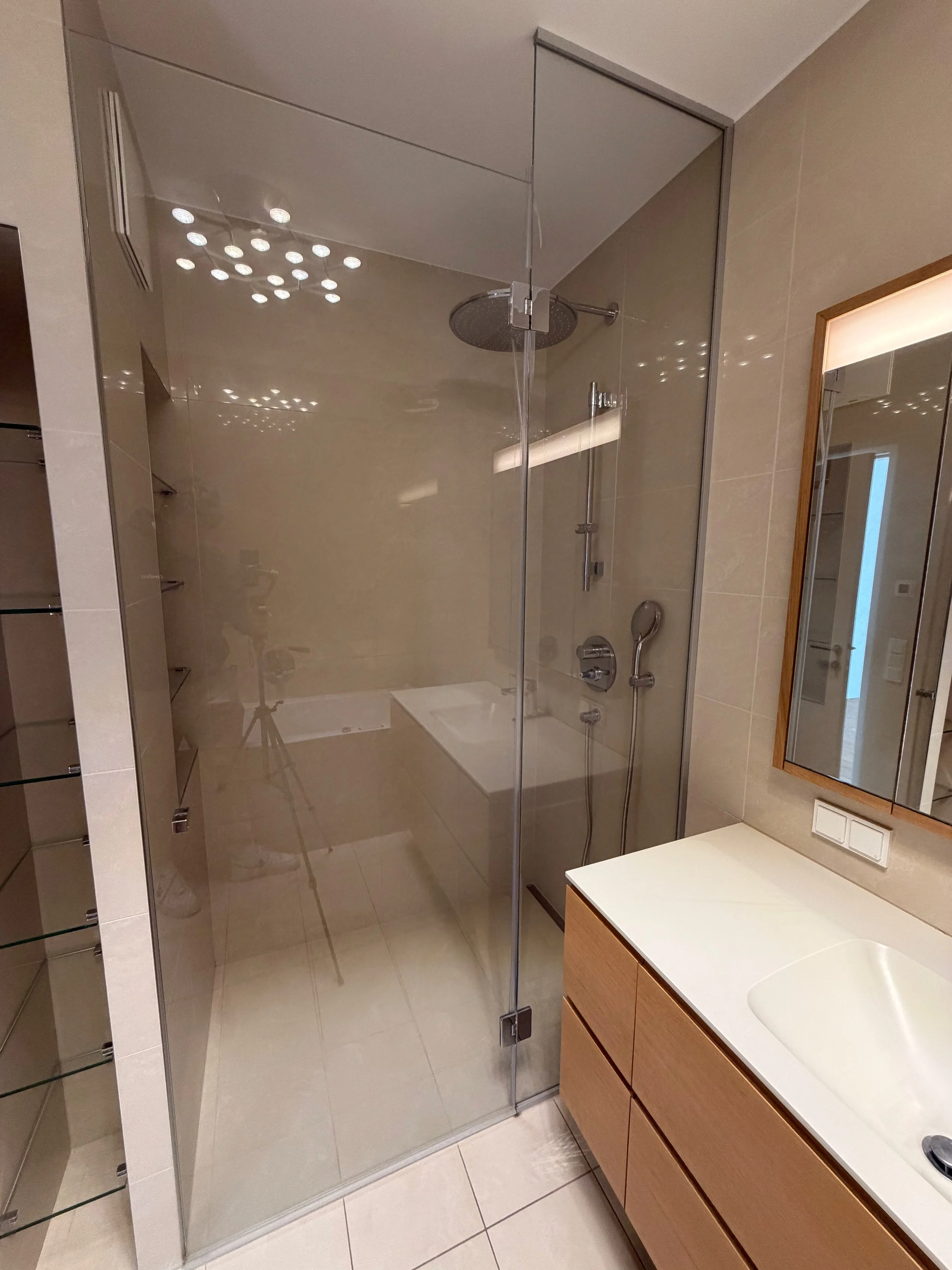 Ein modernes Badezimmer mit einer begehbaren Dusche mit Glastüren, einem Waschtisch mit Spiegel und Holzschränken, beige Wände und Fliesen, sowie einer Deckenleuchte.