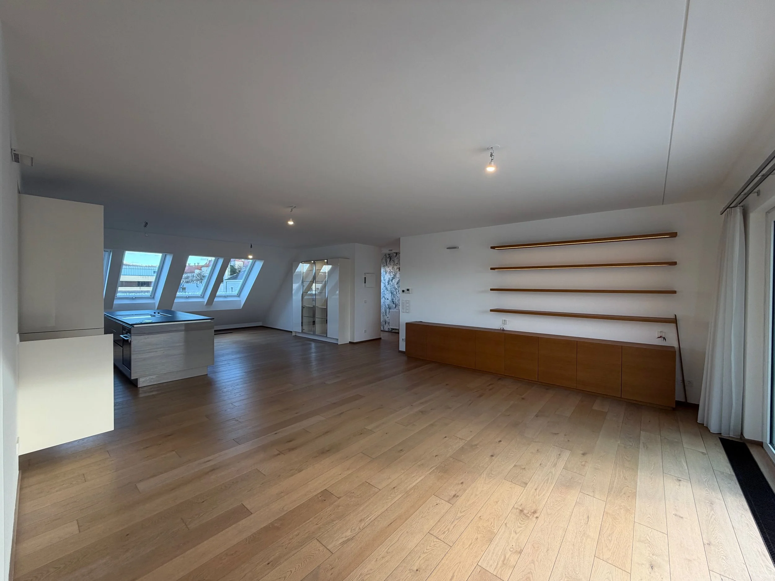 Leeres Wohnzimmer mit Holzfußboden, weißen Wänden, großen Dachfenstern und einer Holzregalwand