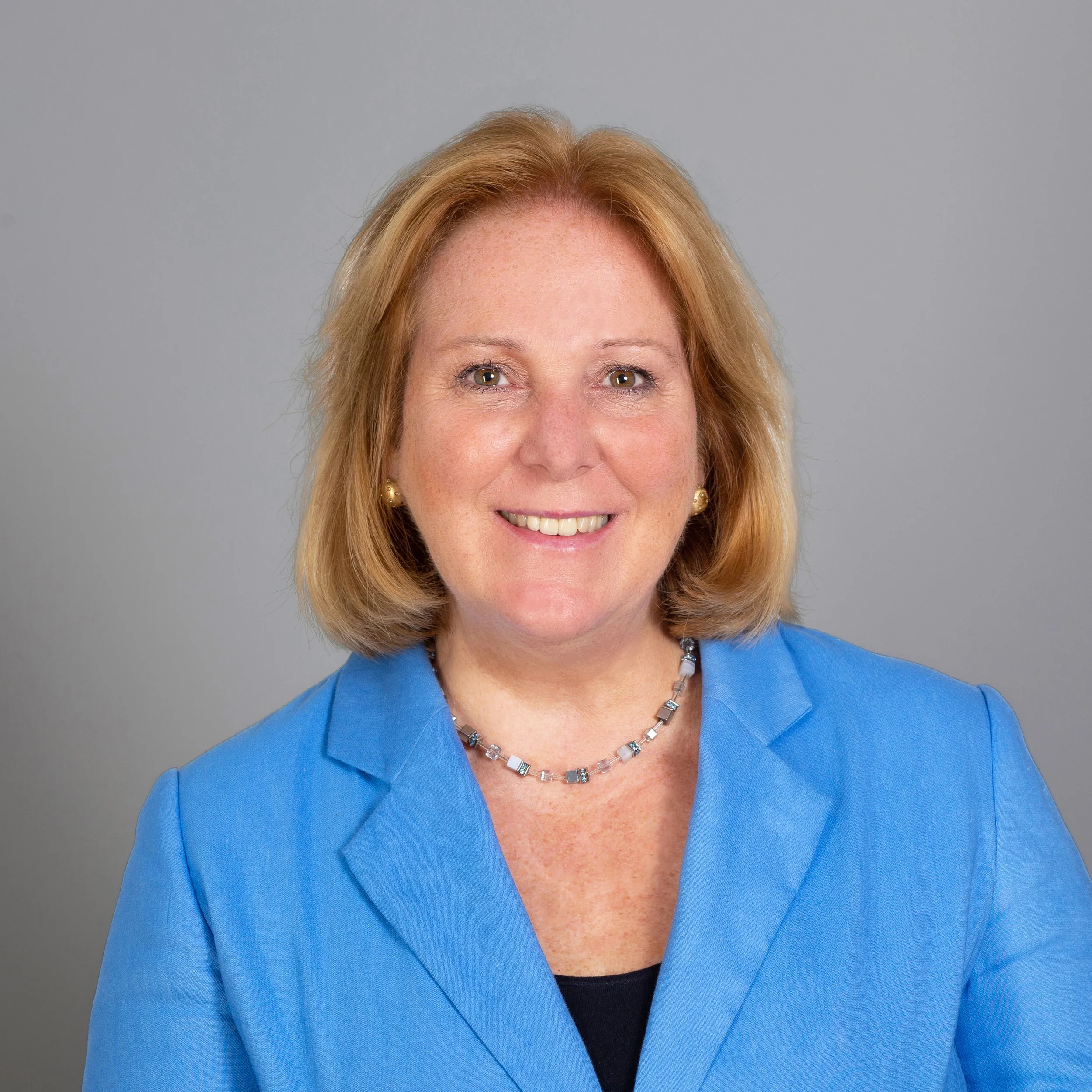 Porträt einer lächelnden Frau mit blonden Haaren, blauer Jacke und Schmuck vor grauem Hintergrund.