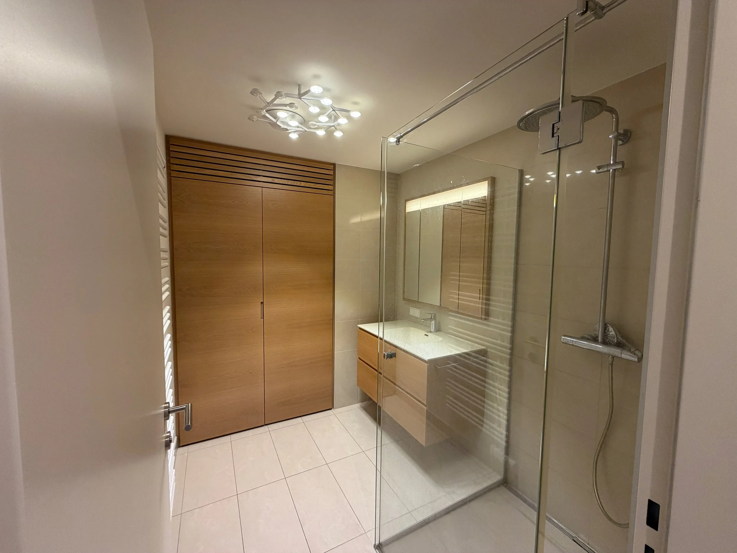 Modernes Badezimmer mit Holzschrank, großem Spiegel, Dusche mit Glaswand und Wandfliesen in neutralen Tönen.