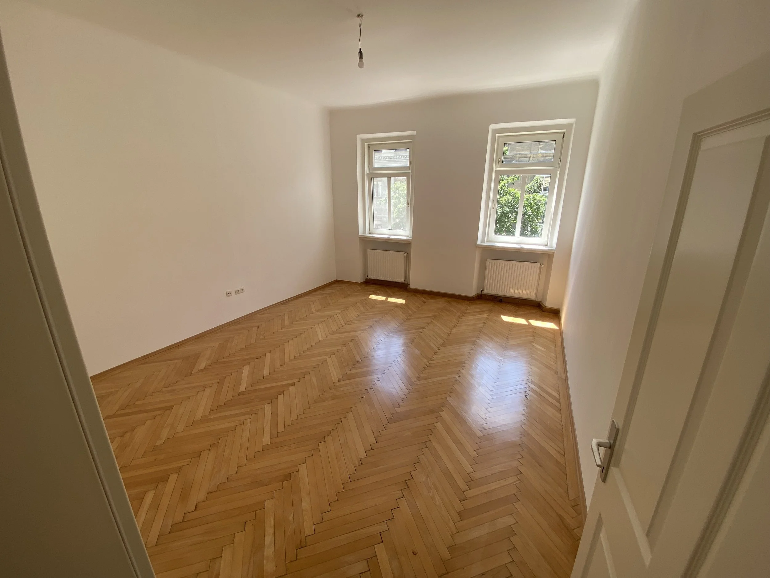 Leeres, helles Zimmer mit zwei großen Fenstern, Holzboden im Fischgrätenmuster, weiße Wände und eine leere Deckenleiste