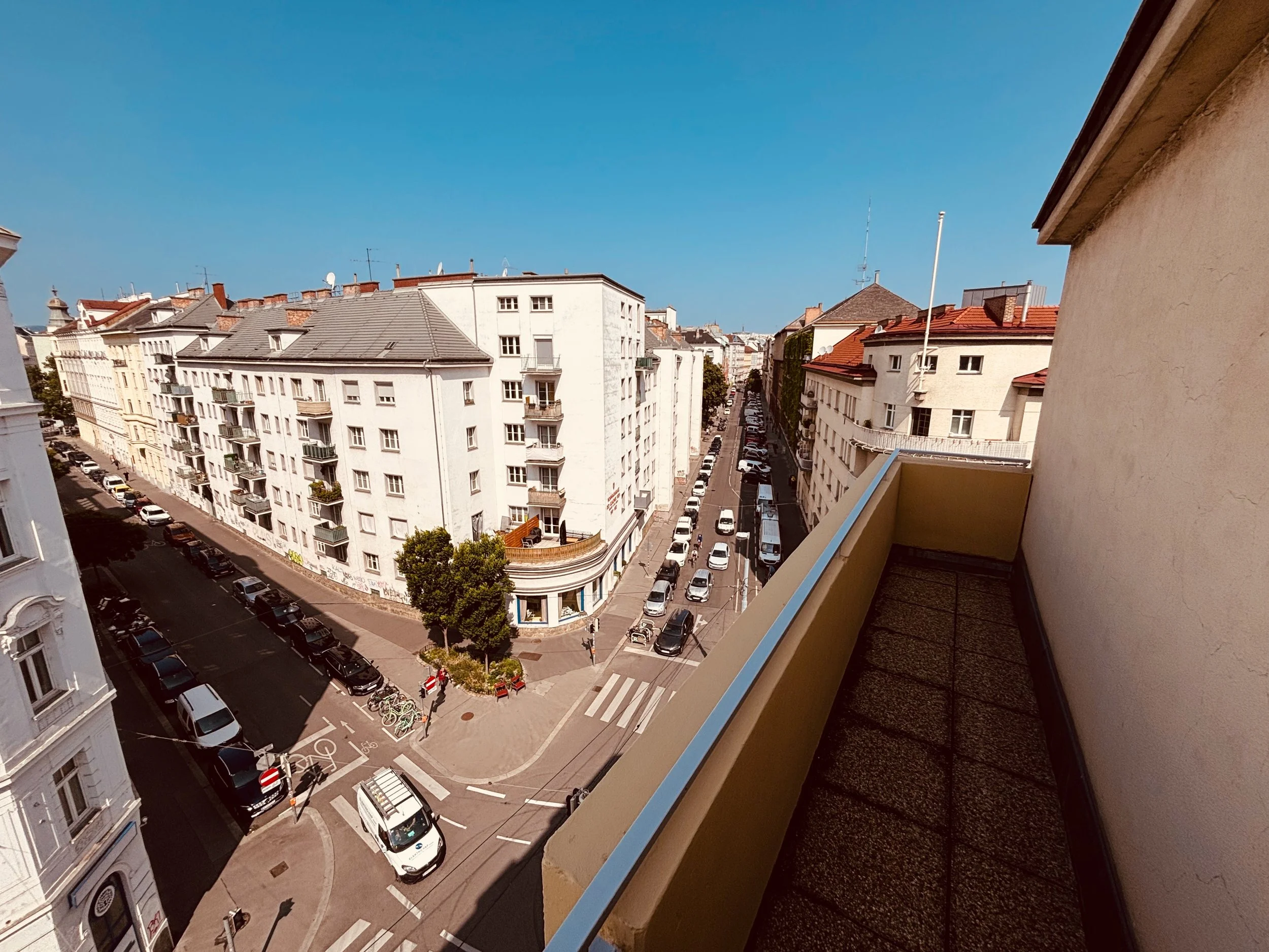 Blick von einem Balkon in eine Stadtstraße mit weißen Gebäuden, Autos und Bäumen bei sonnigem Wetter.
