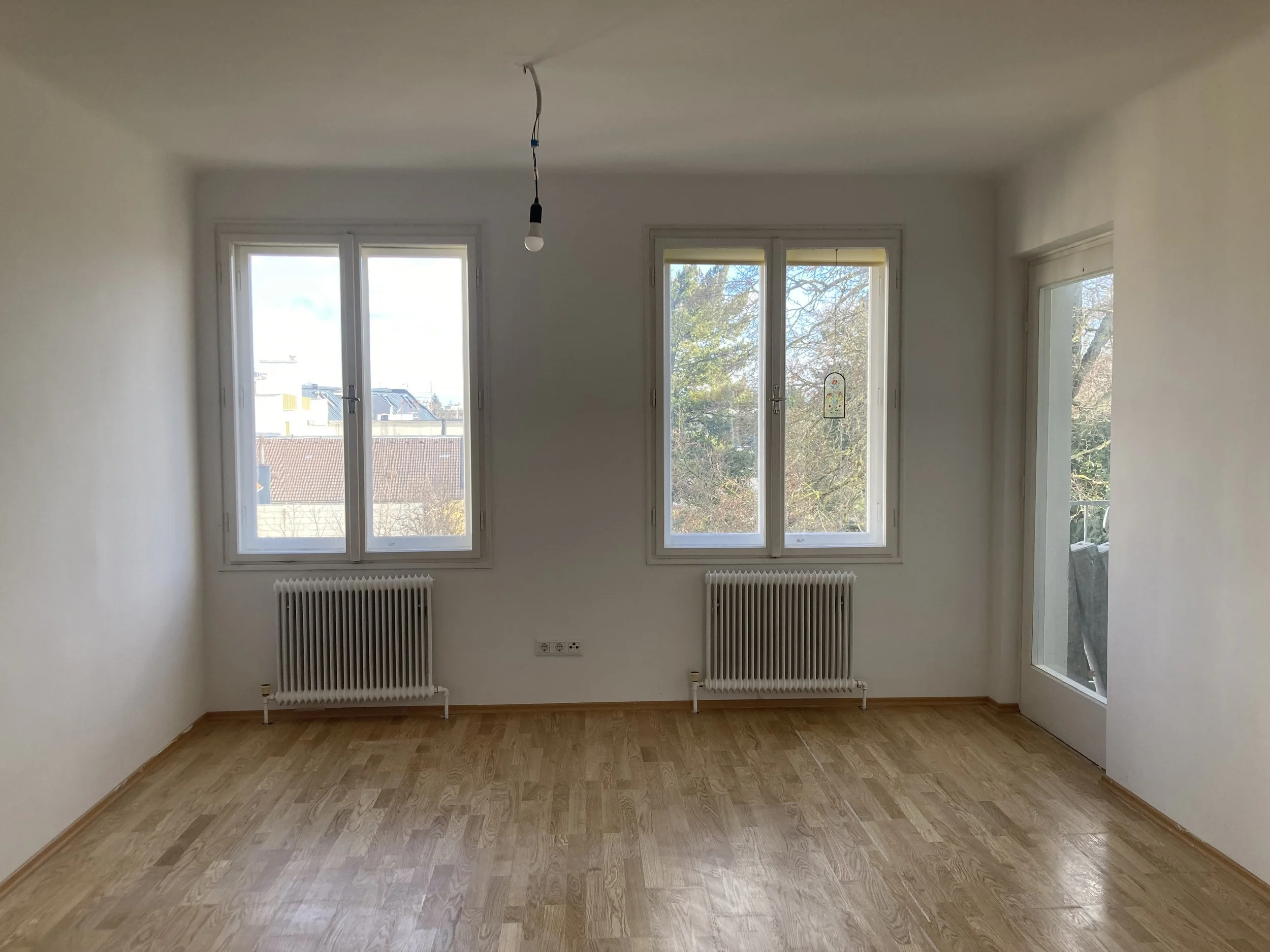 Leeres Wohnzimmer mit drei Fenstern, zwei Heizkörper, Laminatboden und einer frei hängenden Glühbirne