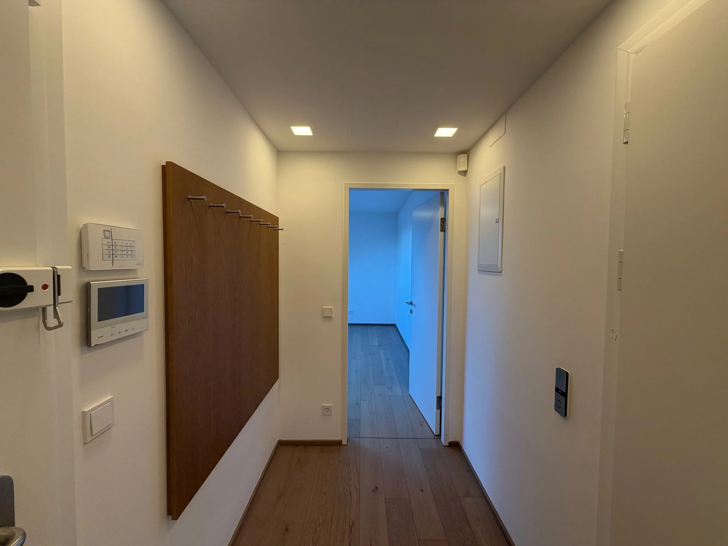 Ein Flur mit weißen Wänden, Holzboden, einer Holztafel mit Haken an der linken Wand, einem weißen Sicherheitssystem und einem Telefon, eine offene Tür führt in einen Raum mit Holzboden, blauer Wand und natürlichem Licht.