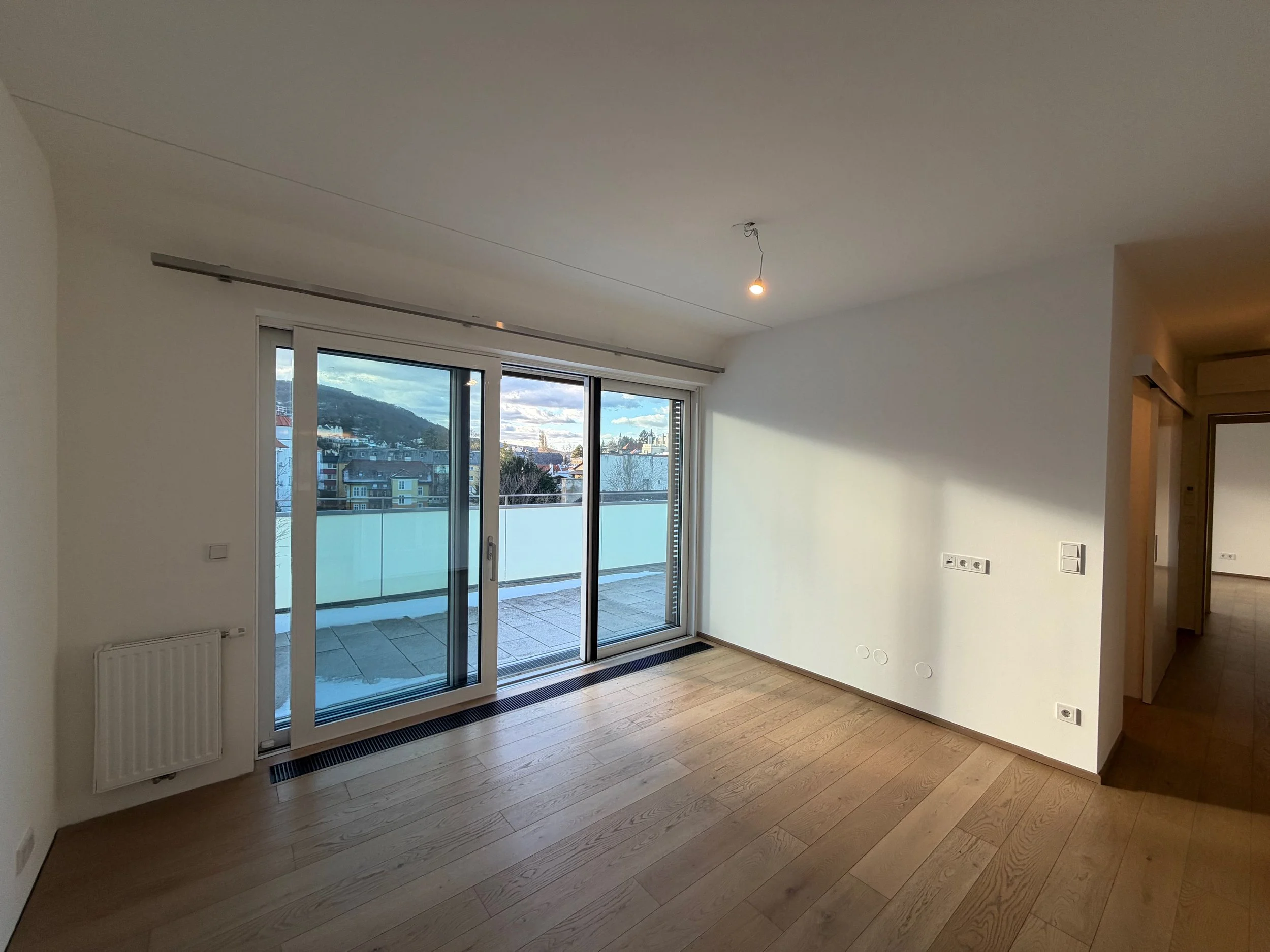 Leeres Wohnzimmer mit Holzfußboden, großen Glasschiebetüren, die auf einen Balkon führen, und einer Kulisse aus Bergen und Gebäuden im Hintergrund.