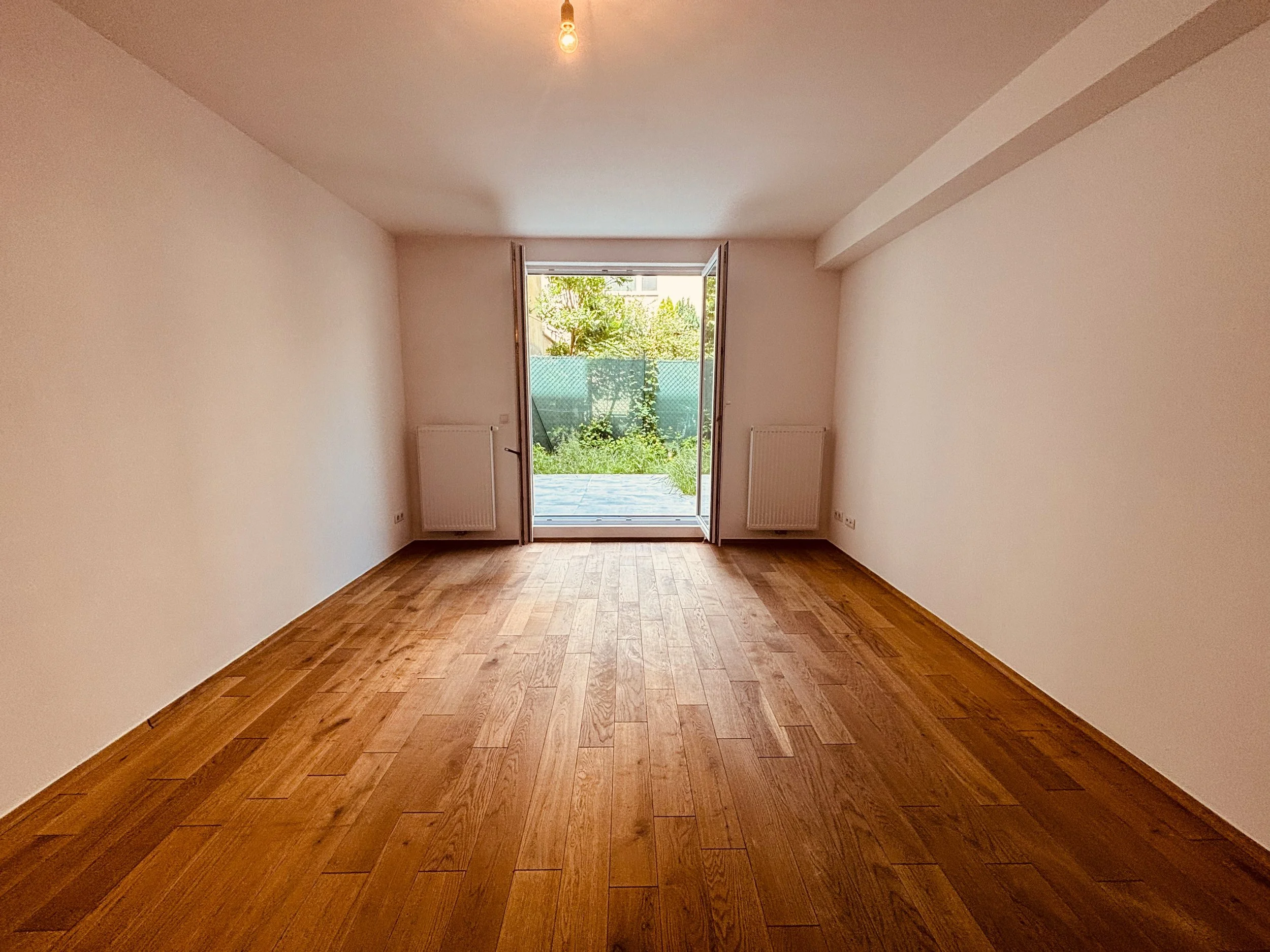 Ein leeres Wohnzimmer mit Holzboden, offener Tür nach draußen auf einen Garten mit Bäumen und Hecke.