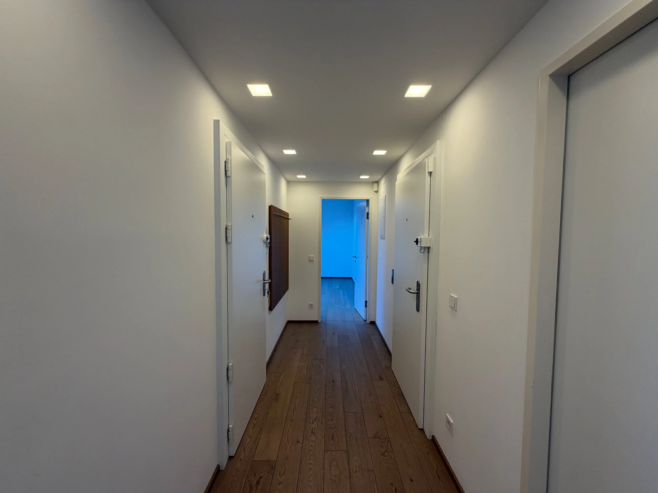 Ein schmaler Flur in einem modernen Gebäude mit weißen Wänden, Holzboden und mehreren weißen Türen, die nach links und rechts führen. Am Ende ist ein blauer Raum sichtbar.