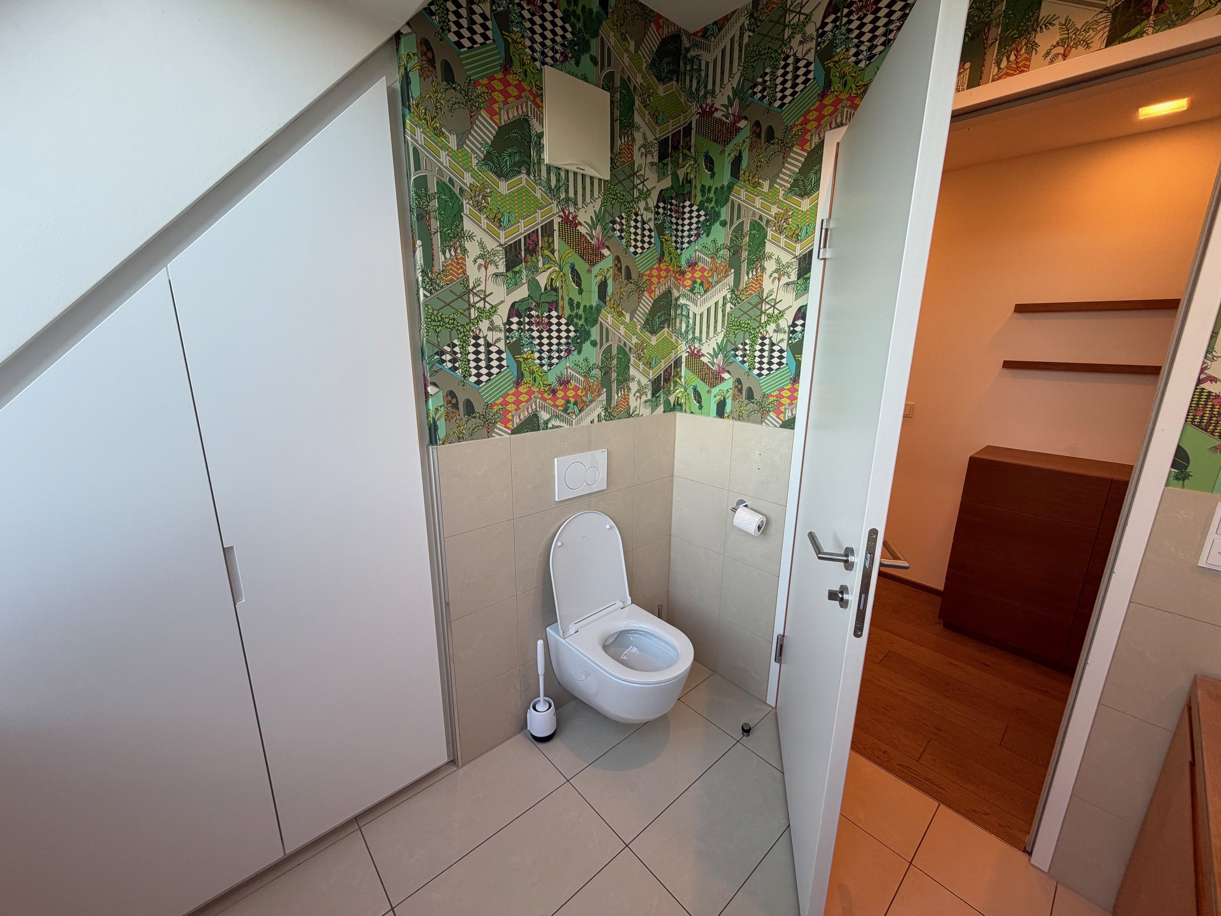 Ein modernes Badezimmer mit einer weißen Toilette und bunten Wandtapete mit einer urbanen, tropischen Landschaft. Es gibt eine weiße Tür und ein geöffnetes Türenpanel, das in einen Holzfußbodenraum führt. Die Wand über der Toilette ist teilweise mit 