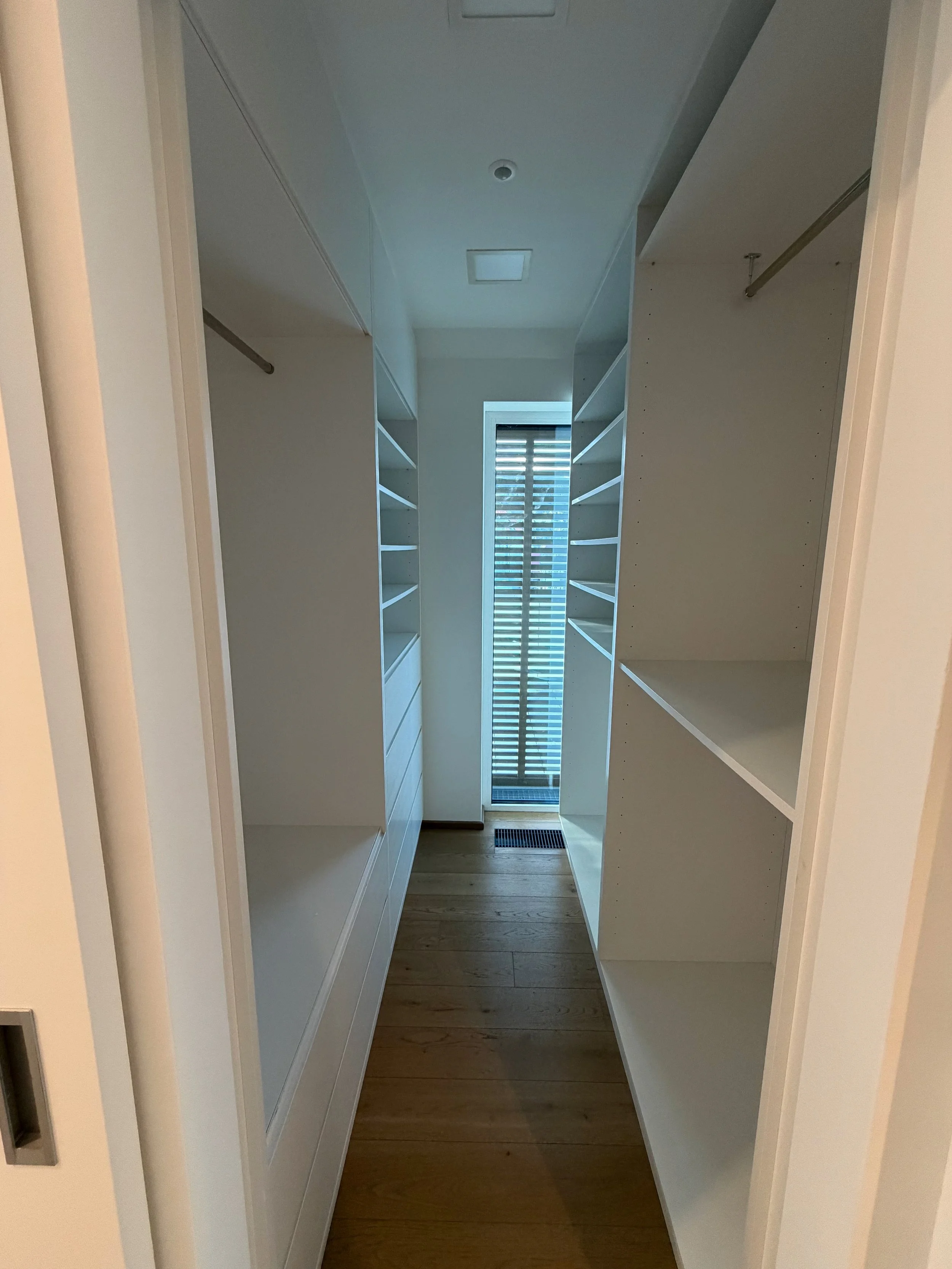 Ein seitlicher Blick in eine leere begehbare Garderobe mit weißen Regalen auf beiden Seiten, Holzboden und einem Fenster am Ende mit Rollos.