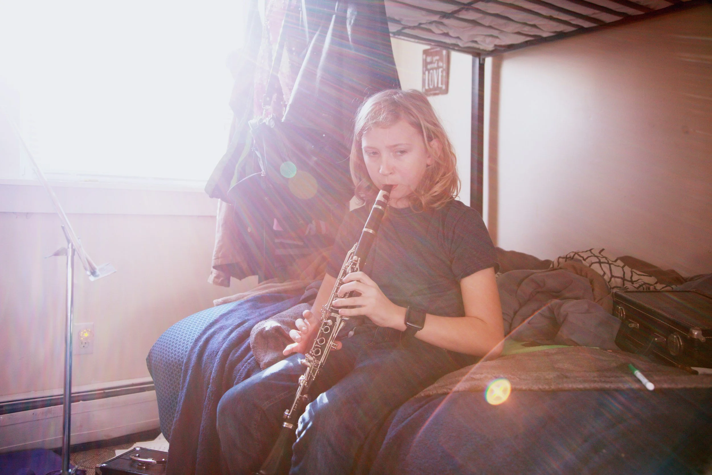 A young girl sitting on a bed playing a clarinet in a room lit by sunlight. There is a black case next to her, and clothing hanging on a rack behind her. A poster with the word 'LOVE' is visible on the wall.
