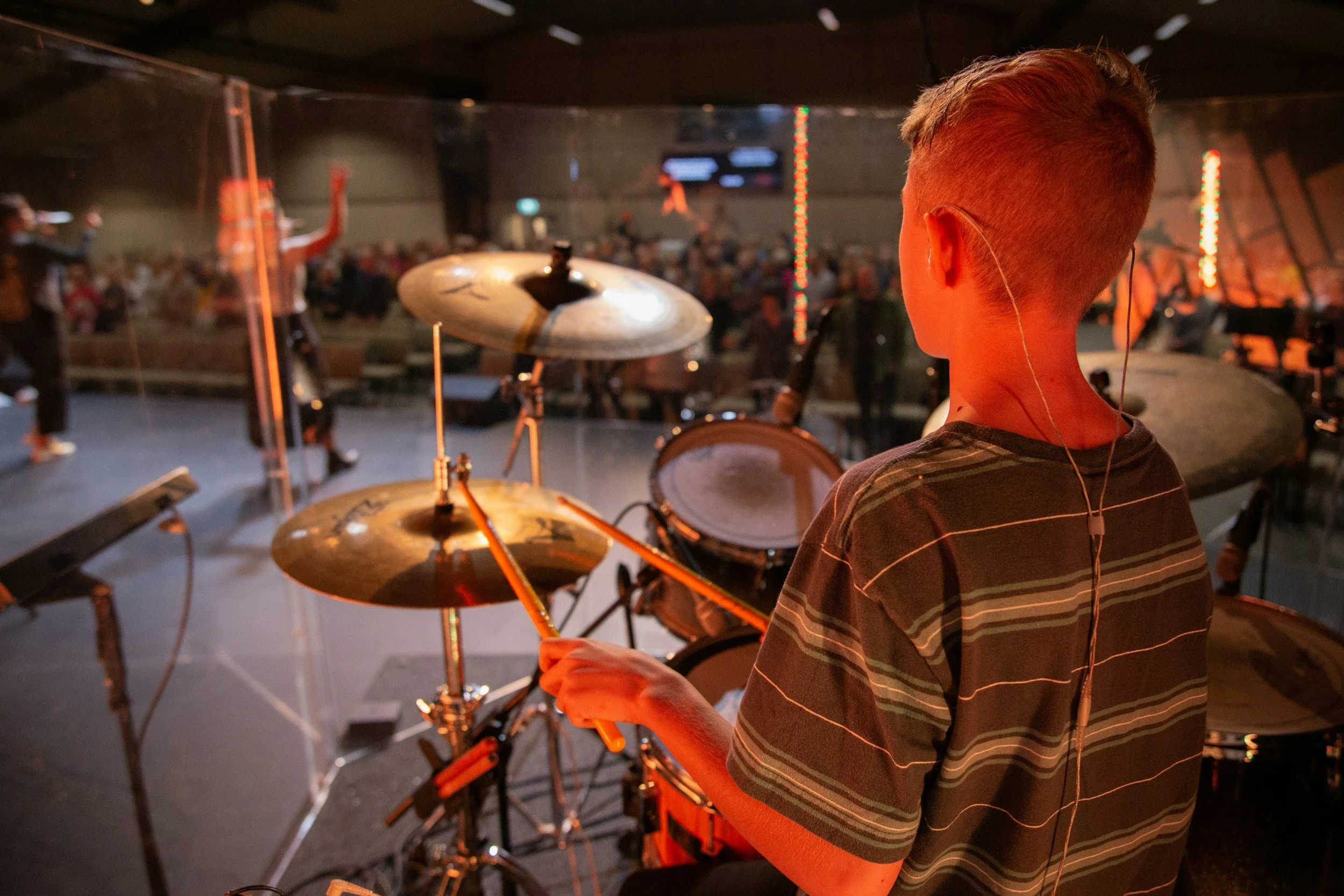 A young person wearing earphones and a striped shirt, seen from the back, is standing backstage at a concert or event, with a drum set and an audience in the background.