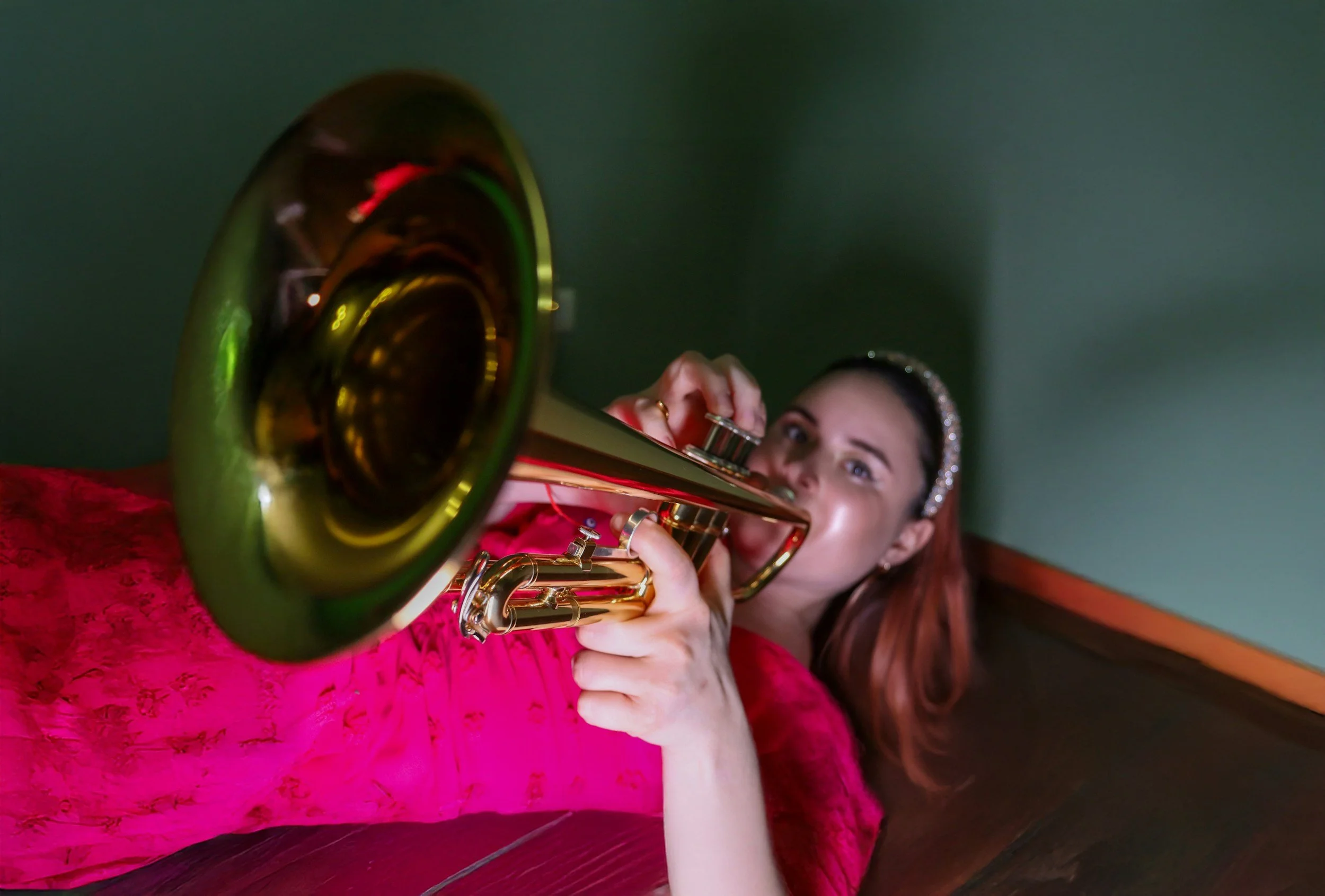 A woman in a pink lace dress playing a gold trumpet in a room with green walls.