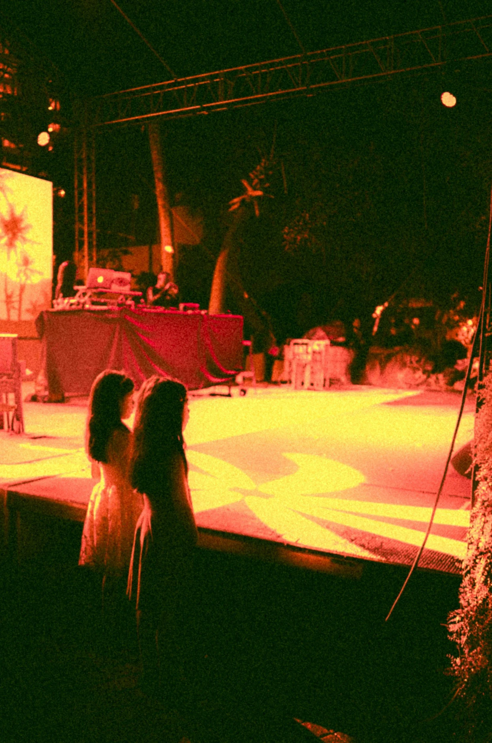Two women in formal dresses standing close together, facing an illuminated outdoor stage with DJ equipment, in a festive environment at night.
