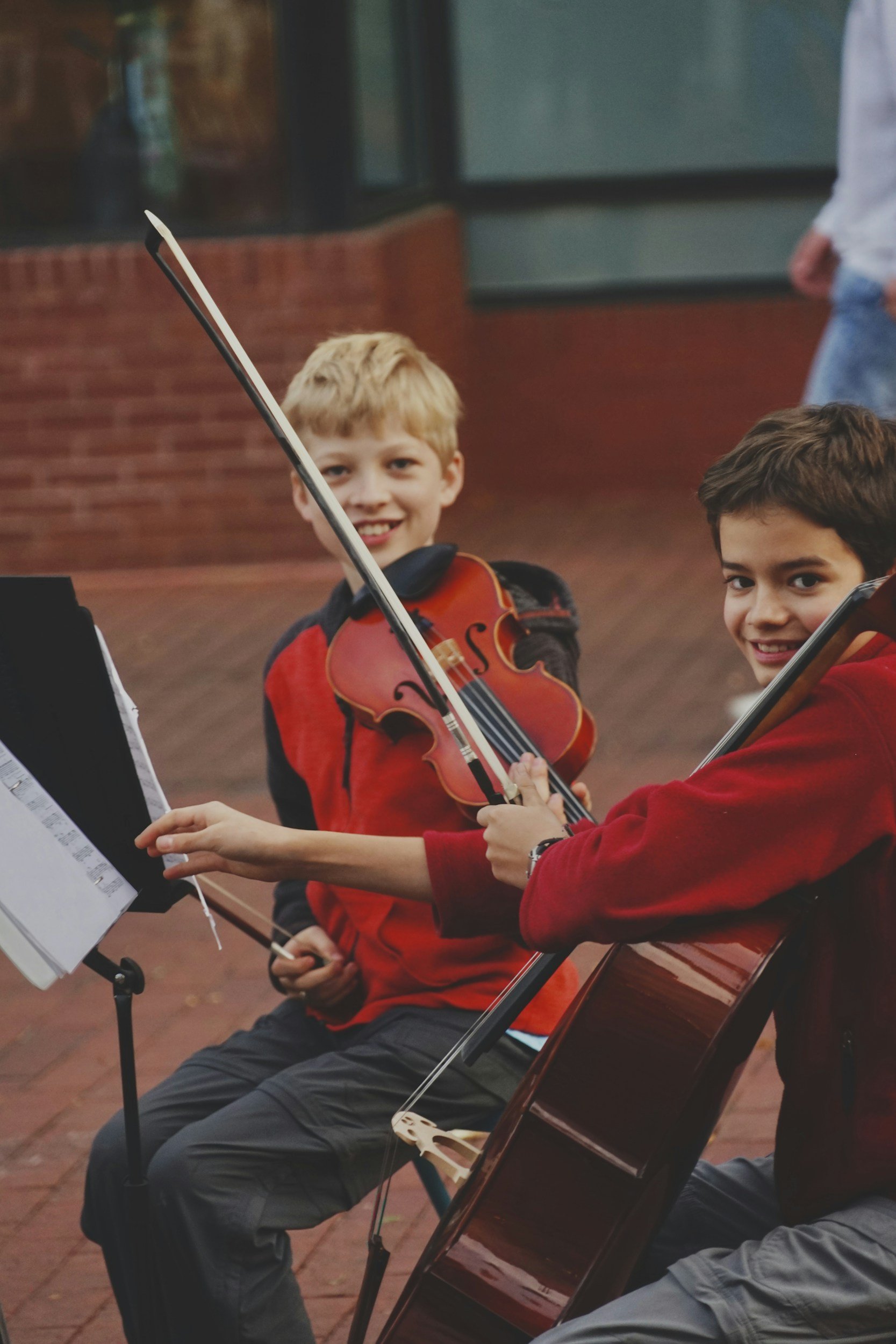 Two young boys playing violins during a music practice, one with blond hair wearing a red hoodie, the other with dark hair also in a red hoodie, sitting on chairs outdoors against a brick wall.