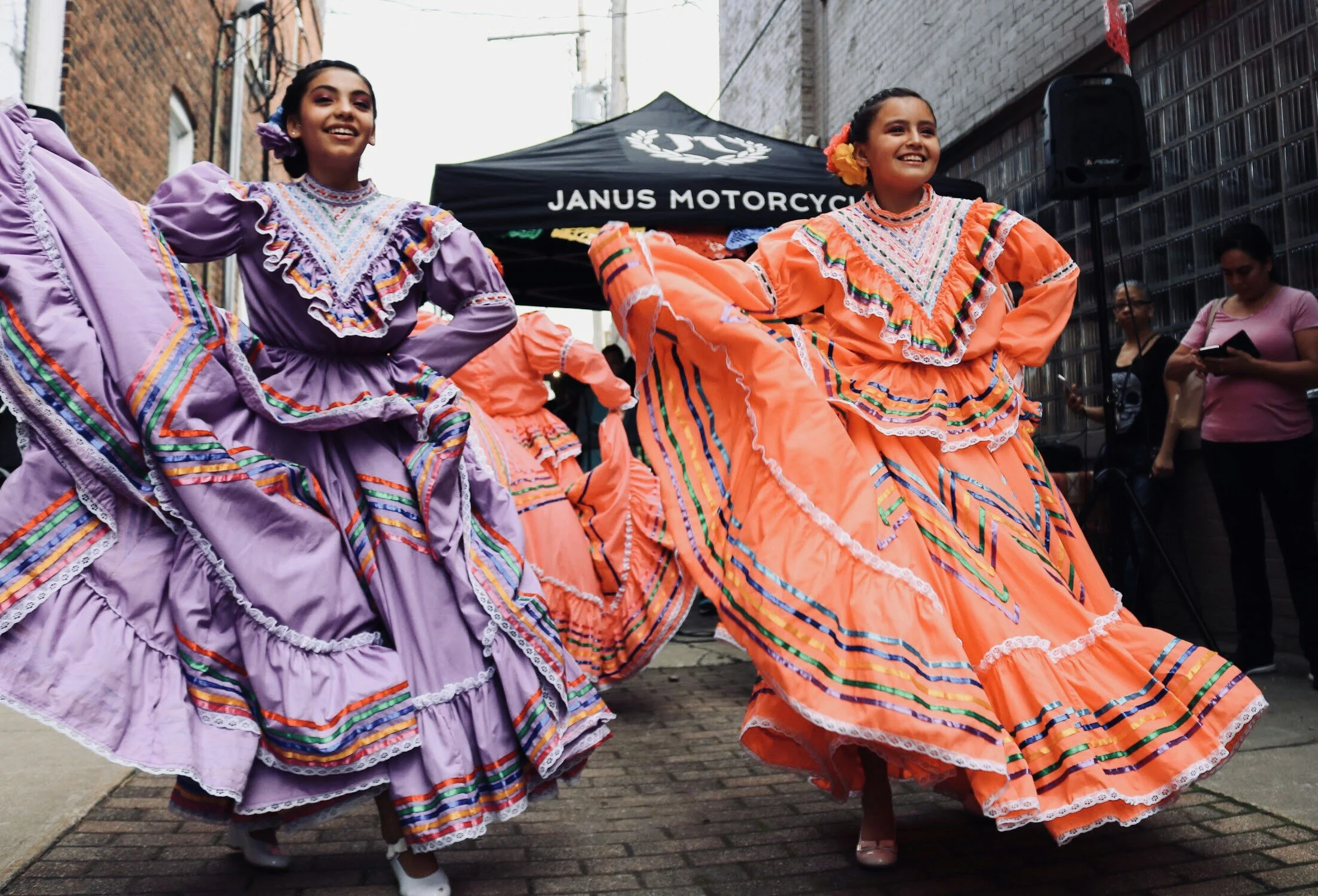 Two women in colorful traditional Mexican dresses dancing on a street during a cultural celebration, with spectators in the background.