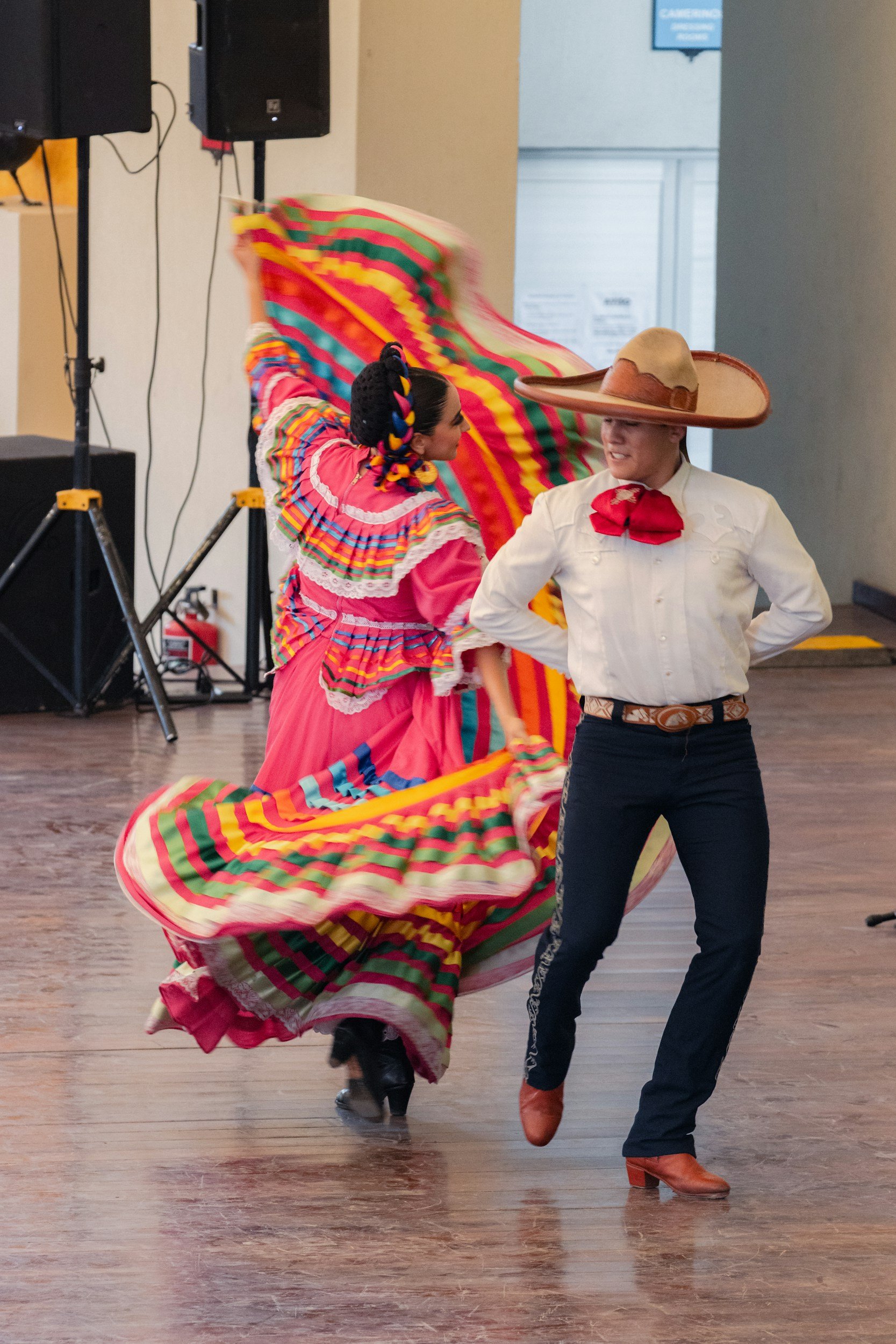 A woman in traditional Mexican attire dancing with a man wearing a giant sombrero, colorful dress, and cowboy boots in an indoor setting.