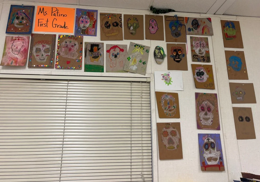 Collection of children's artwork, mainly skull-shaped faces, displayed on a wall with a sign indicating it is Ms. Patino's First Grade class.