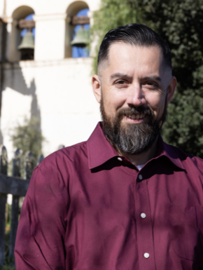A man with dark hair and a beard wearing a maroon dress shirt, posing outdoors with a historic building and trees in the background.