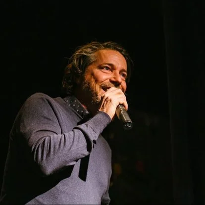 A man with long hair and a beard speaking into a microphone on stage, wearing a gray sweater.