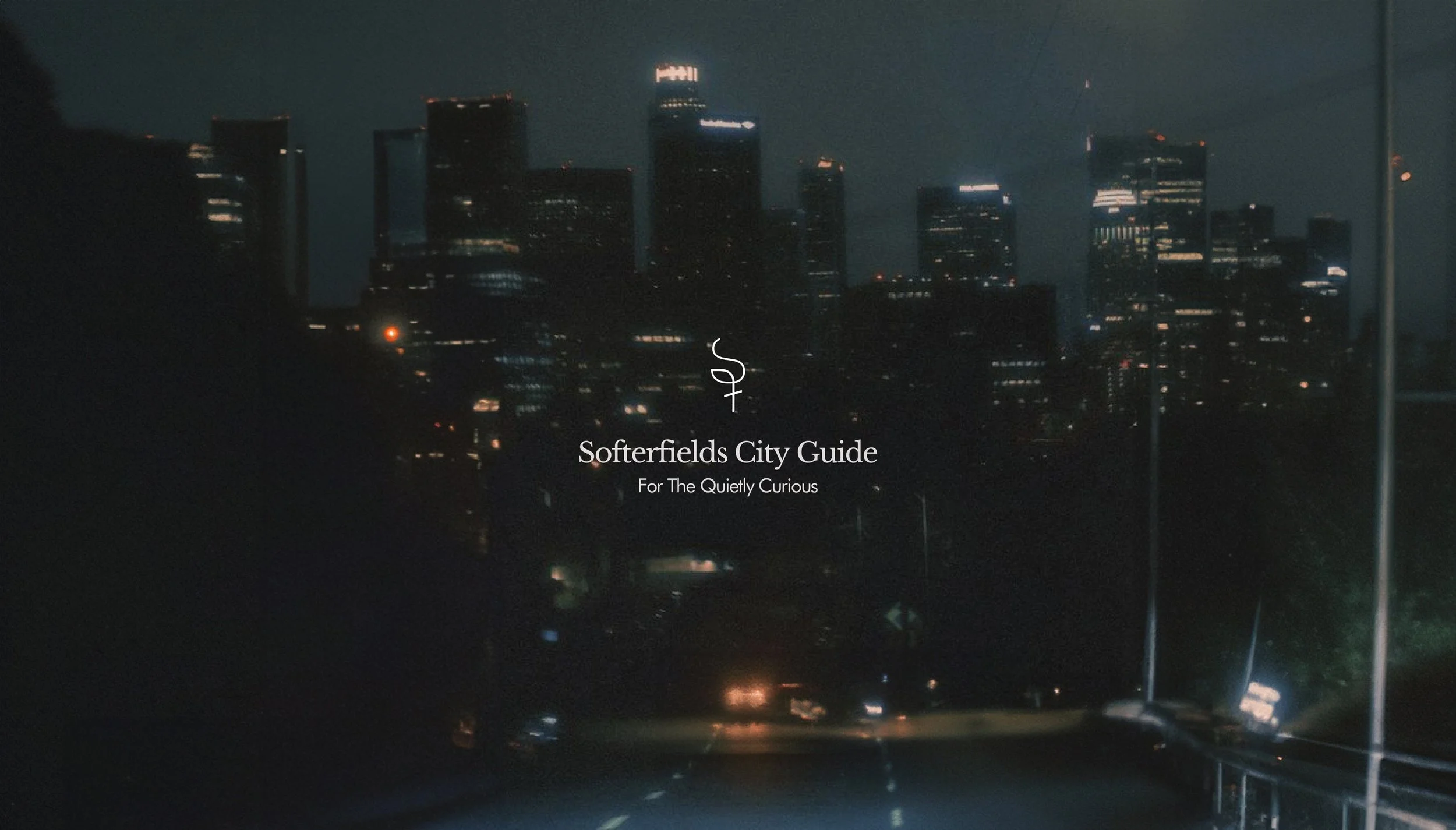 Night view of a city skyline with tall illuminated buildings and a dark sky, overlaid with white text that reads 'Softerfields City Guide' and a tagline 'For The Quietly Curious'.