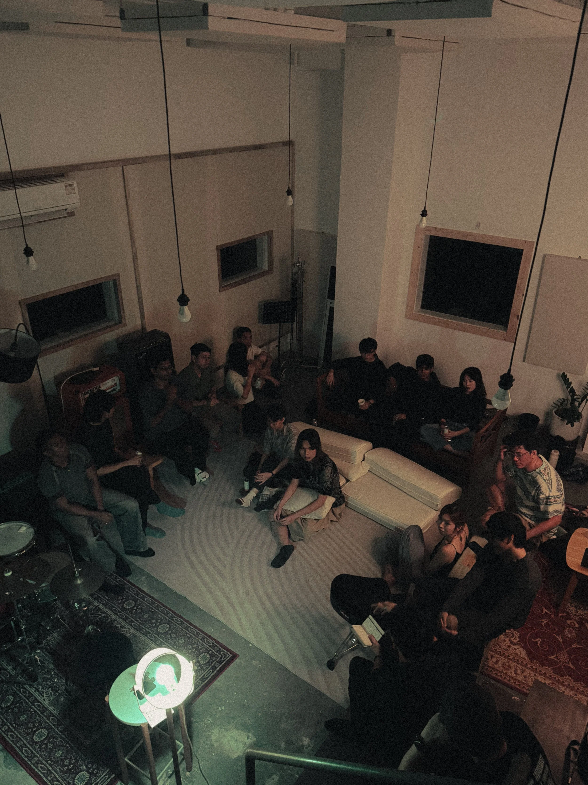 A group of people gathered in a cozy living room, sitting on sofas and chairs, with some holding drinks, engaged in a social gathering or meeting, under low lighting with hanging light bulbs.
