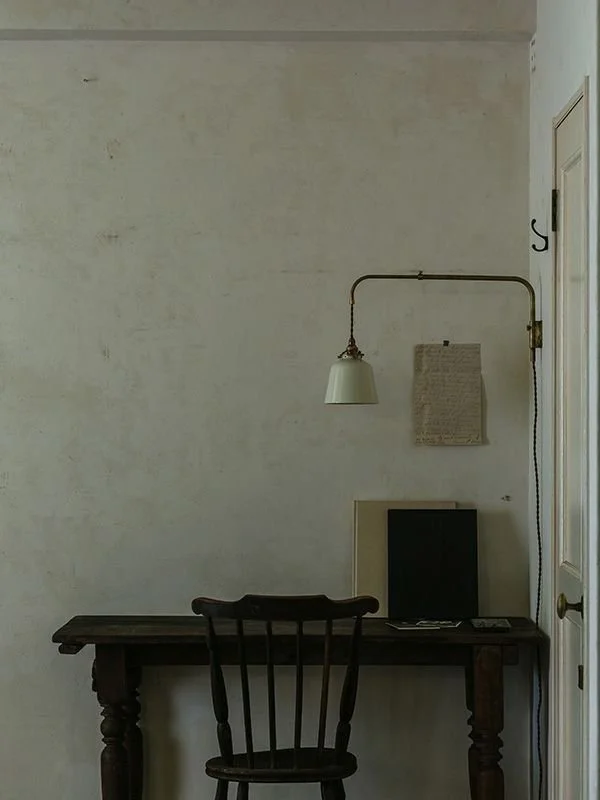 A rustic wooden desk with a black chair in front, an old computer monitor, and a beige radiator or heater behind them. A wall-mounted lamp with a brass pipe and white shade extends above the desk. A piece of paper or fabric is taped to the wall next to the lamp, and the wall is painted white with some visible wear and stains.