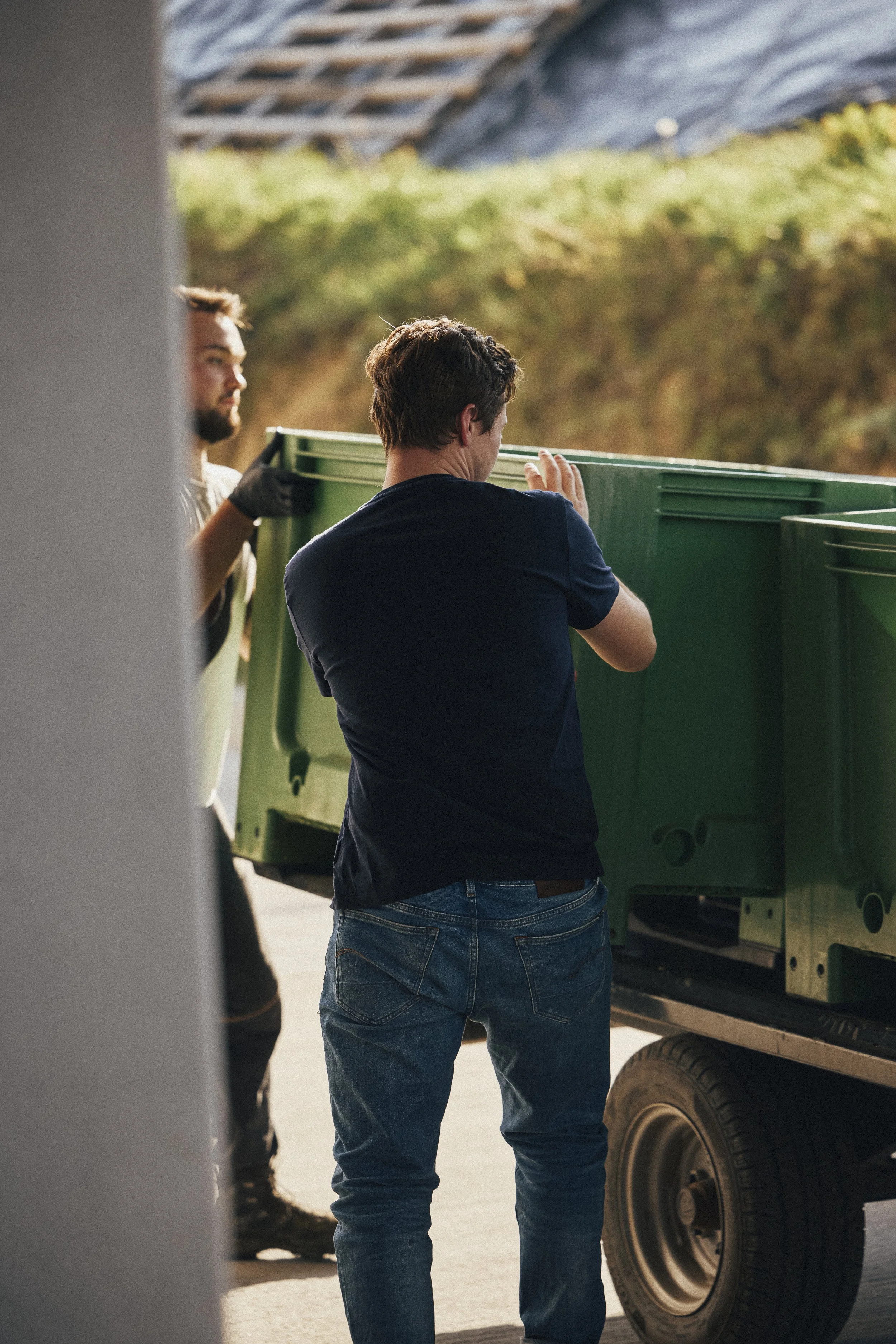 Zwei Männer heben einen grünen Müllcontainer an einen LKW. Der Hintergrund zeigt eine grüne Wiese und eine steile Felswand.