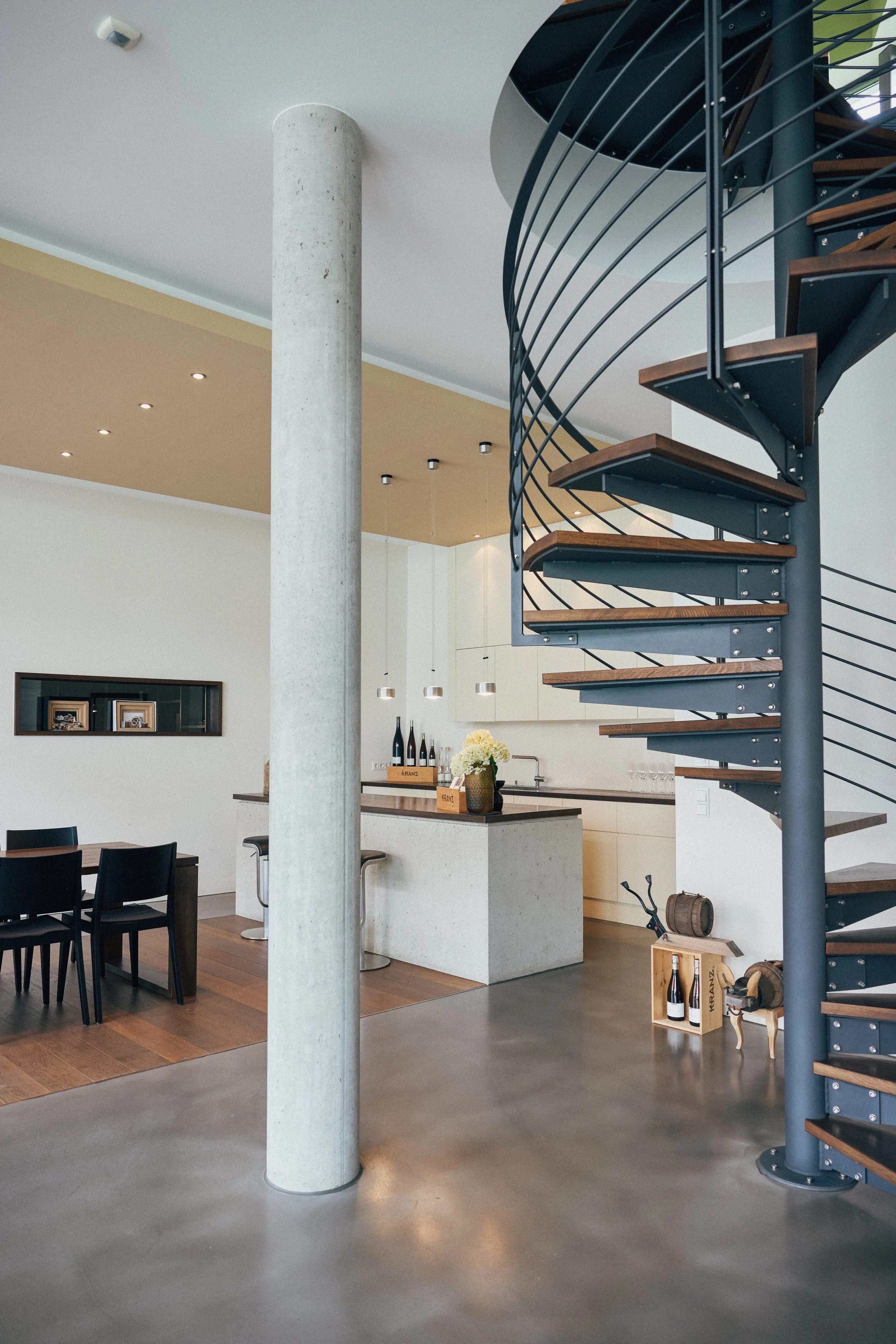 Innenaufnahme einer modernen Vinothek mit Weinverkauf in der Pfalz. Spiraltreppe, Küchenbereich mit Blumen und Weinflaschen, dunklen Esstisch mit schwarzen Stühlen, helle Wände und Holzdekorationen.