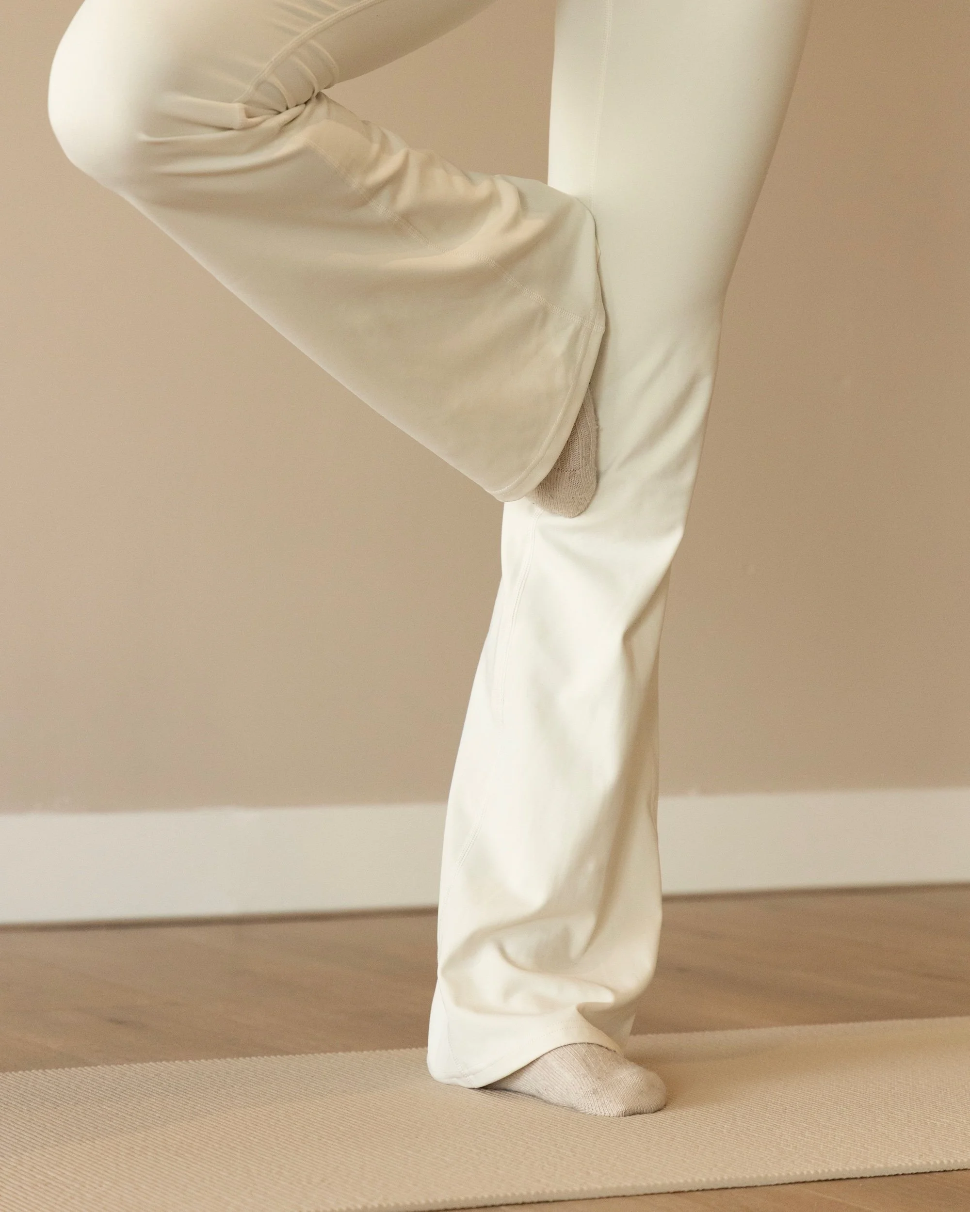 Person practicing yoga or stretching, standing on a mat with one leg raised and bent at the knee, wearing beige or cream-colored pants and socks.