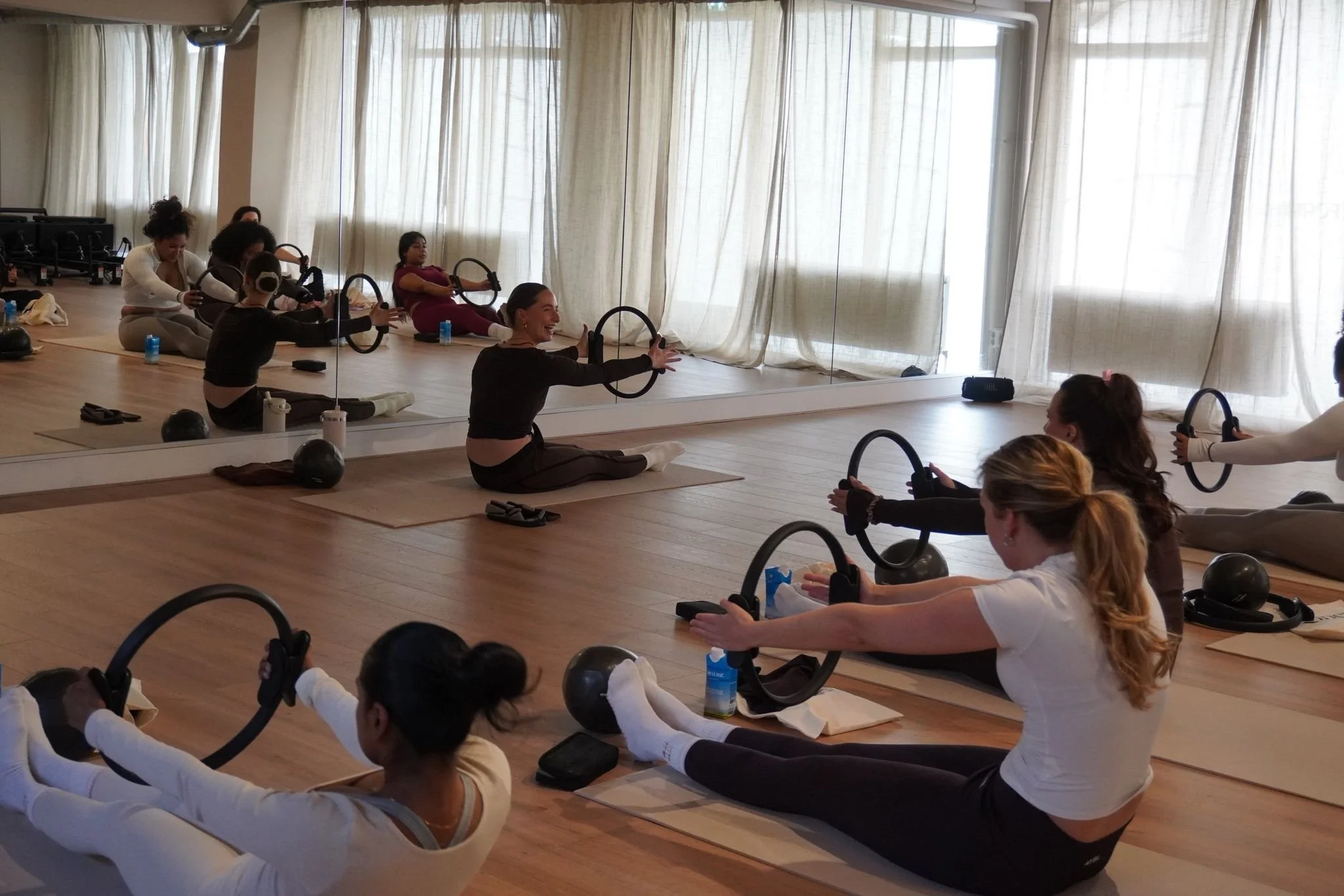 Women participate in a Pilates class using Pilates rings in a spacious studio with a mirror wall and large windows with curtains.