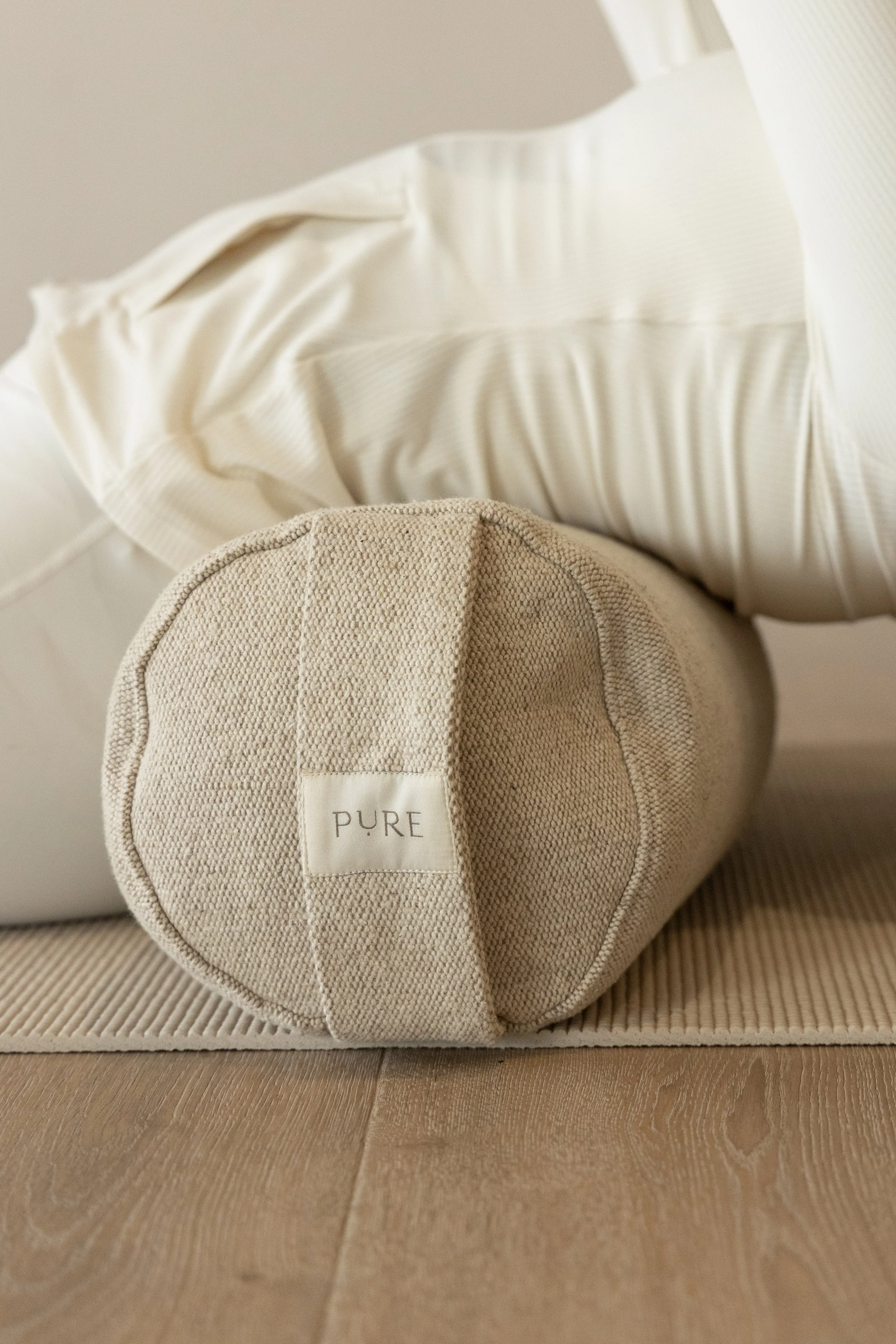 A beige fabric round bolster pillow with a attached white tag labeled 'PURE' resting on a wooden floor next to a cream-colored bed with white sheets and a cream pillow.