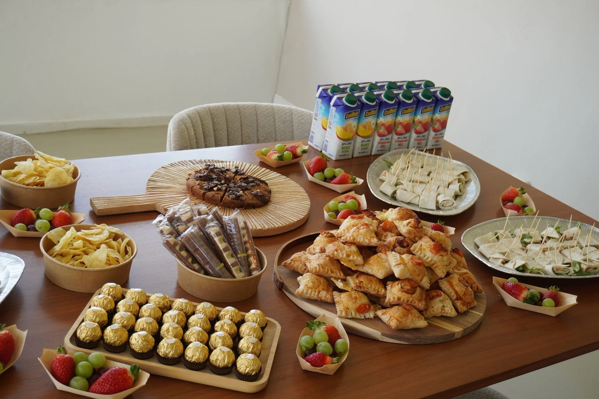 Table set with assorted snacks and beverages including chocolate chip cookies, fruit, mini sandwiches, pastry puffs, candies, and juice boxes.