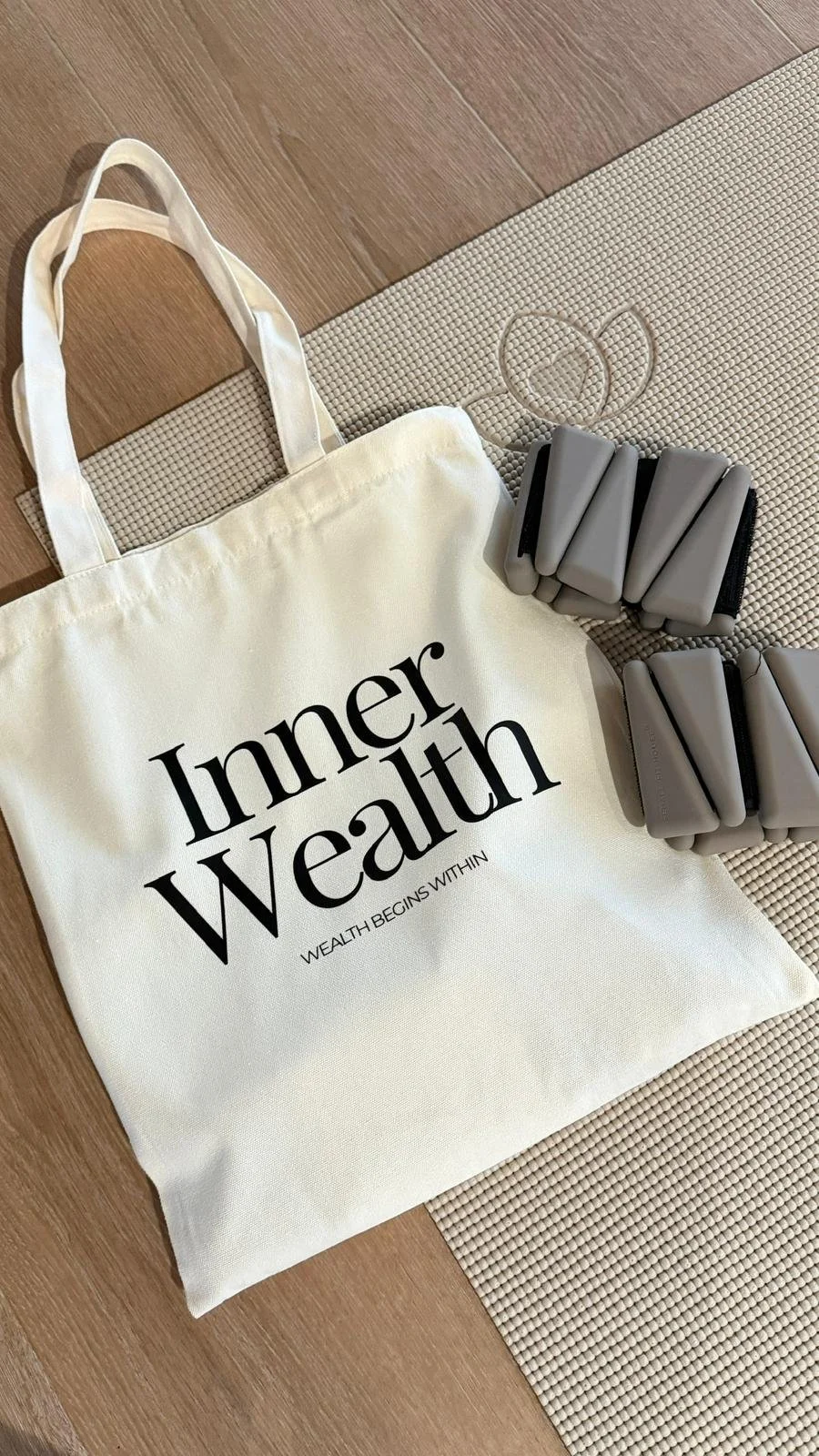 A white canvas tote bag with black text reading 'Inner Wealth' and 'Wealth begins within,' placed on a wooden floor next to a yoga mat. Nearby are two sets of gray foam massage balls.