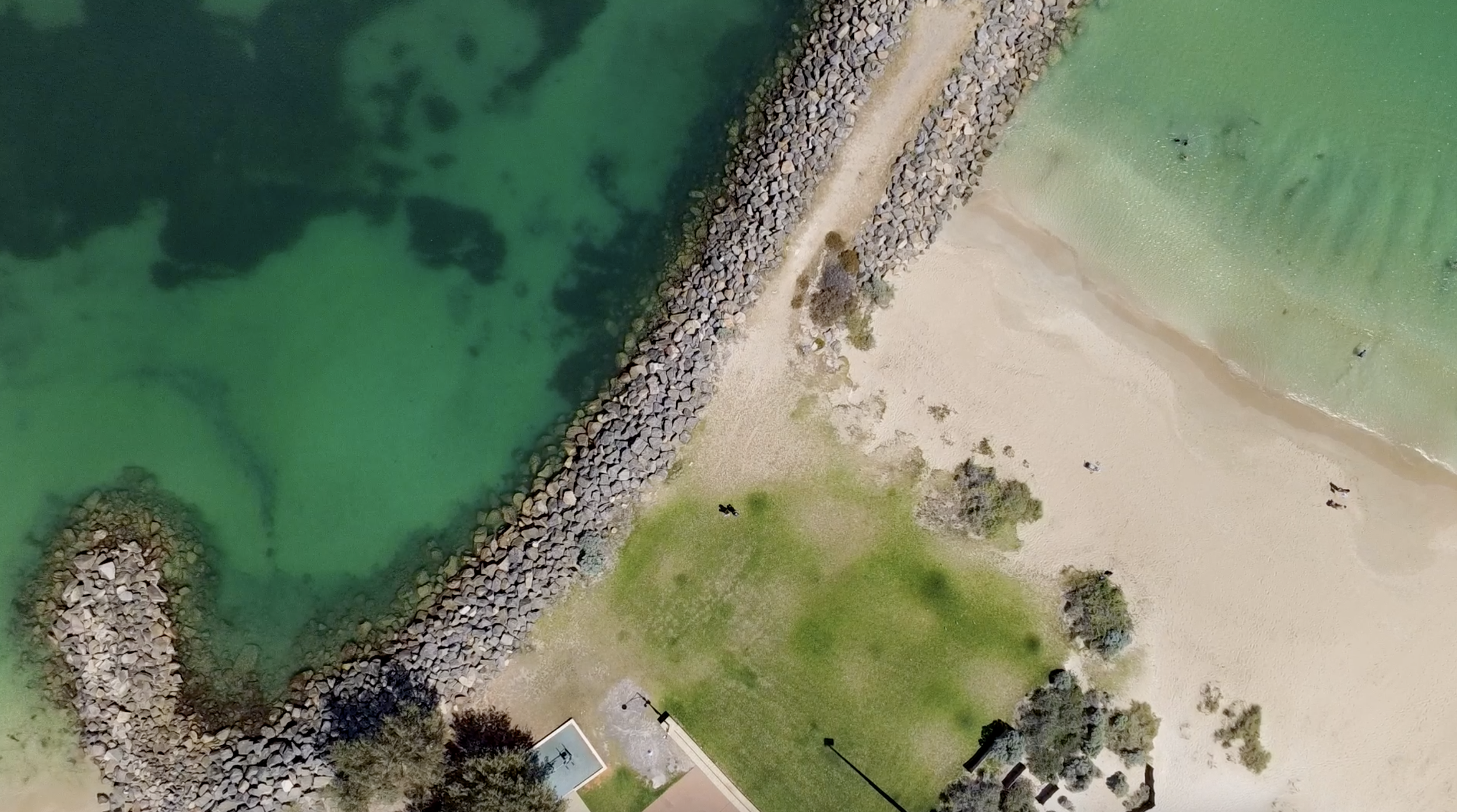 An aerial view of a beach with a rocky breakwater on the left side, green water on the left and right, sandy shoreline, and some small trees and grass area with lamp posts.