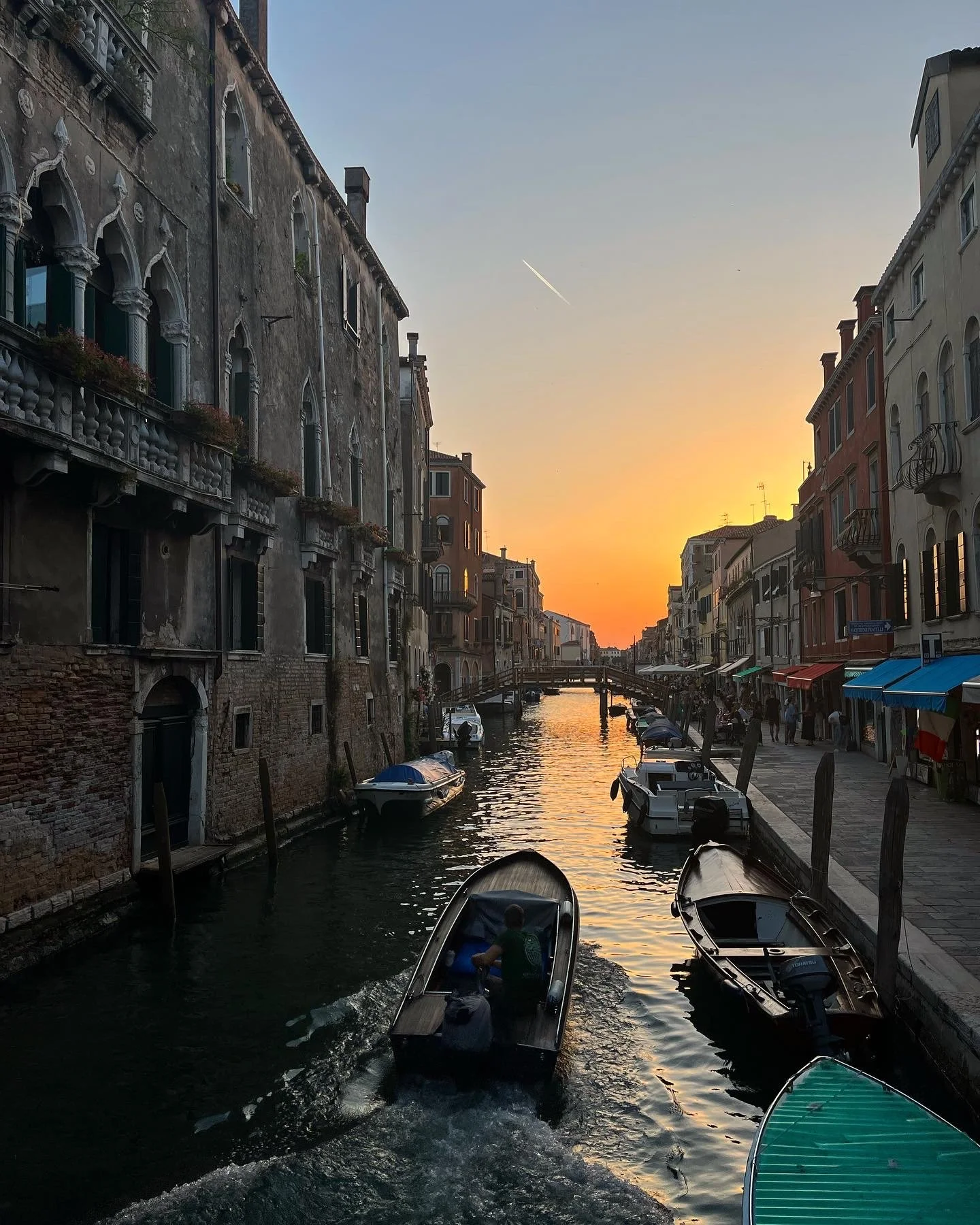 Sunset over a canal in Venice, Italy with boats, historic buildings, and a bridge in the background.