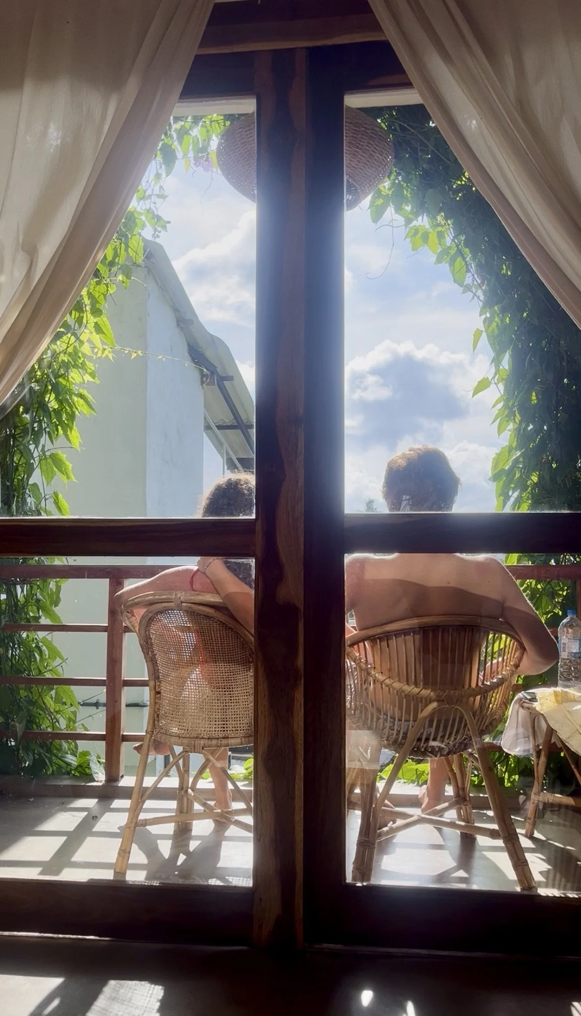 Two people sitting on a balcony, viewed through glass doors, with sunny weather and partly cloudy sky.