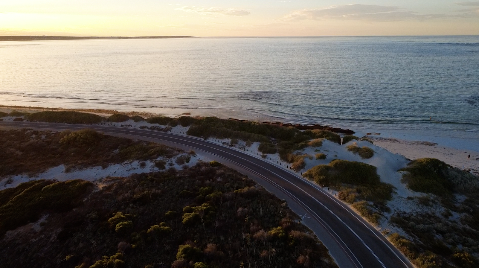 An aerial view of a coastal road winding through sandy dunes and green vegetation, leading to a calm ocean under a partly cloudy sky during sunset.