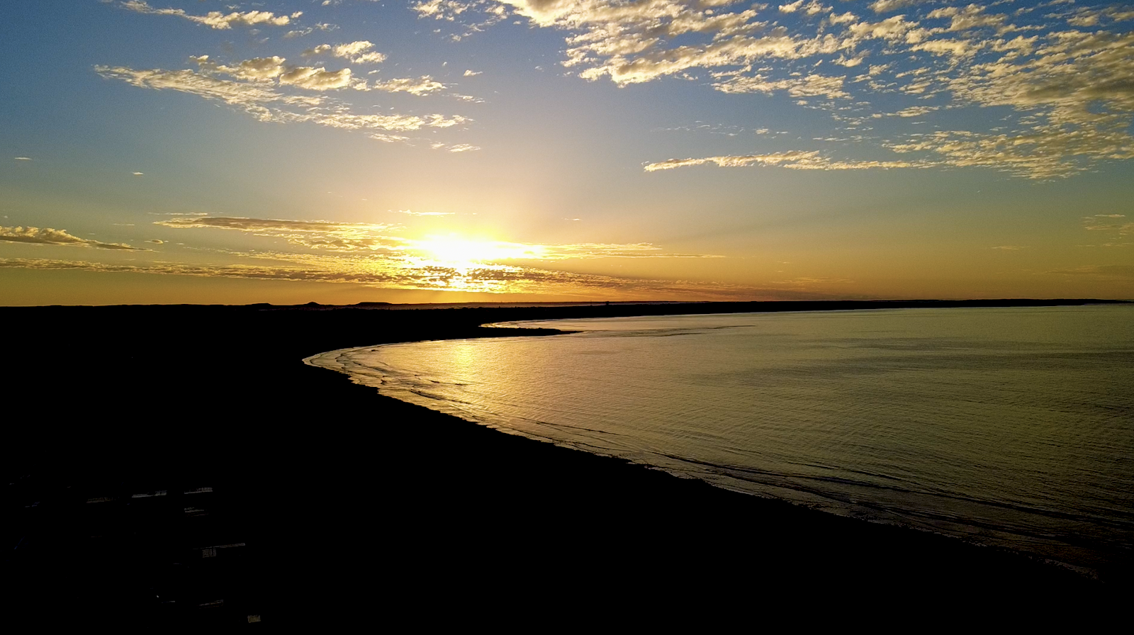 Sunset over a calm beach with a curved shoreline, reflecting the golden light on the water, and scattered clouds in the sky.