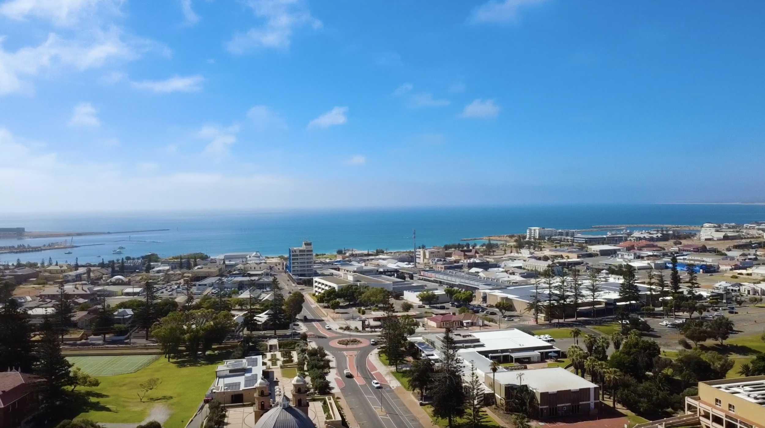 Aerial view of a coastal city with buildings, roads, trees, a park, and the ocean in the background.