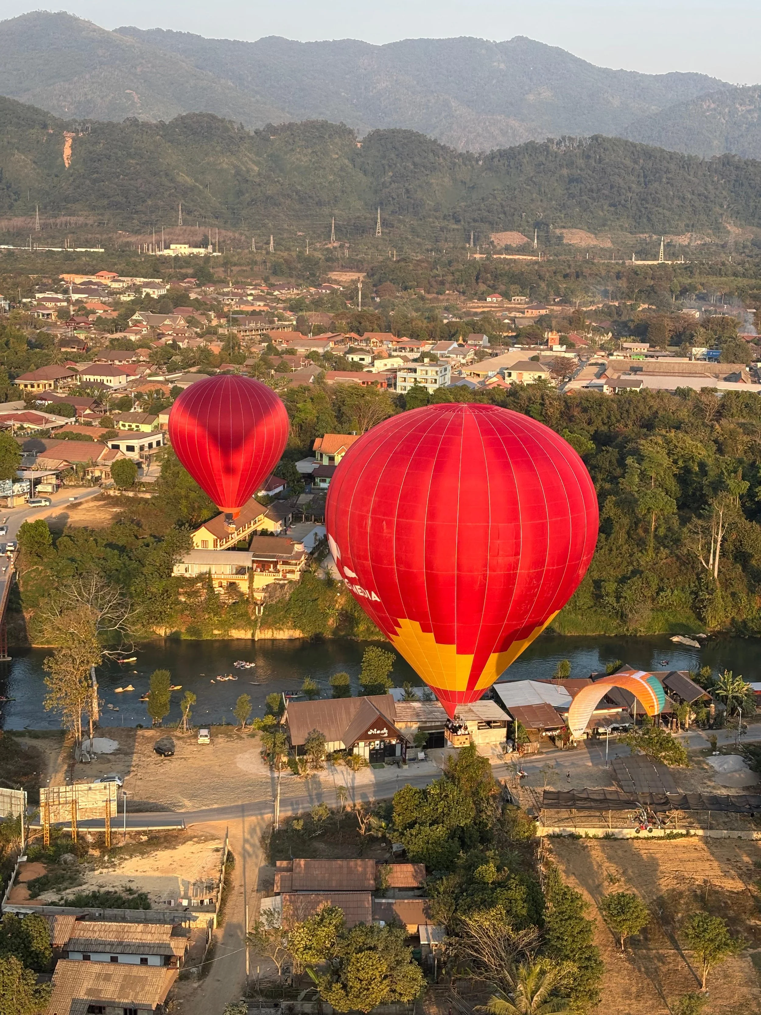 Two red hot air balloons floating over a town near a river, with mountains in the background during sunset.