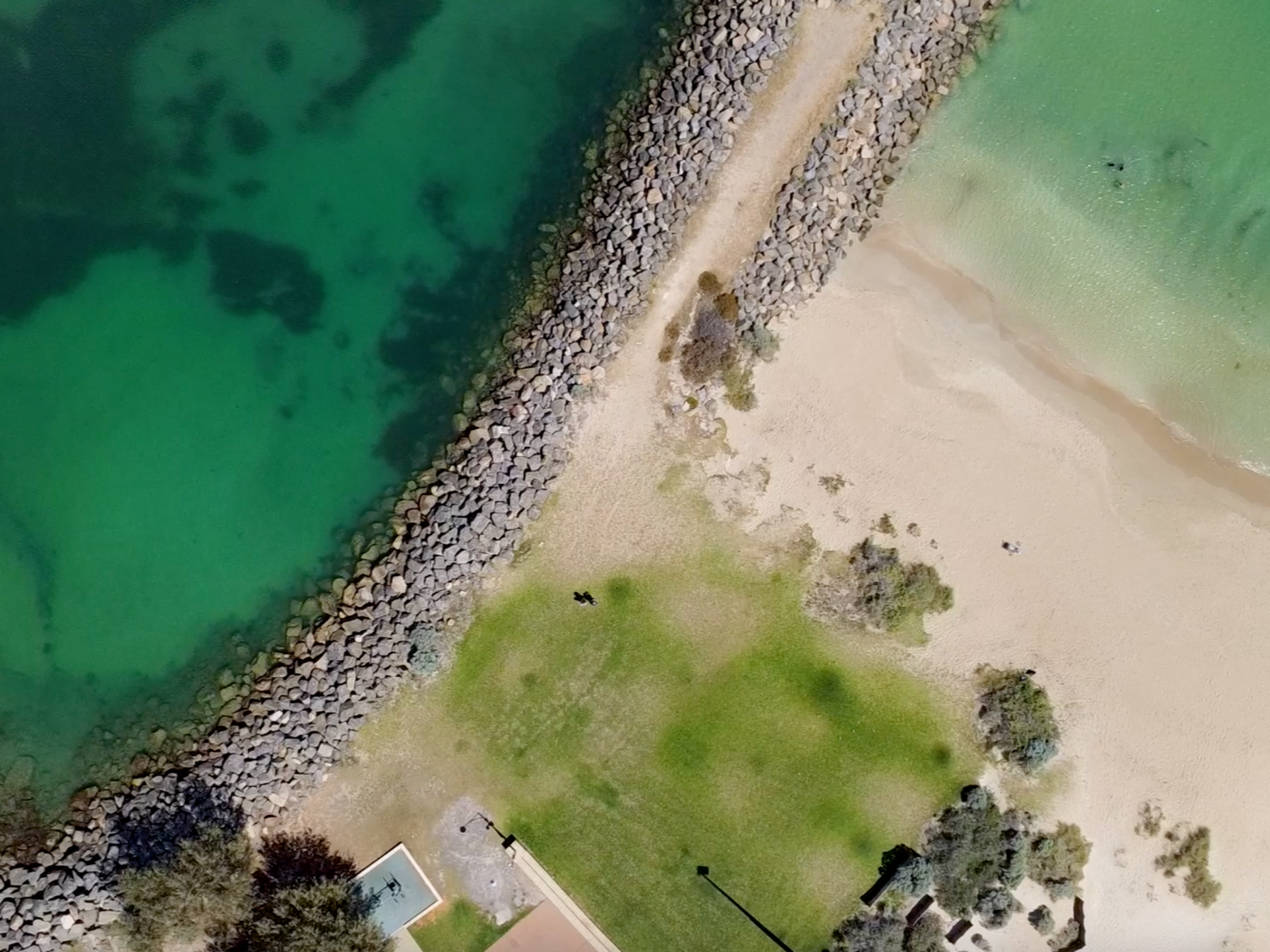 An aerial view of a beach with a shoreline, green water, a sandy beach, a grassy area with trees, and a rocky breakwater.