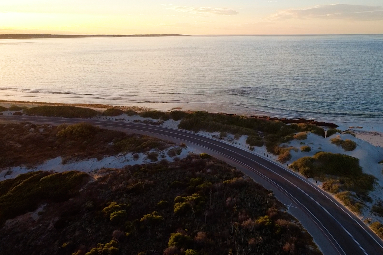 Aerial view of coastal highway winding through sand dunes with vegetation, overlooking the ocean at sunset.