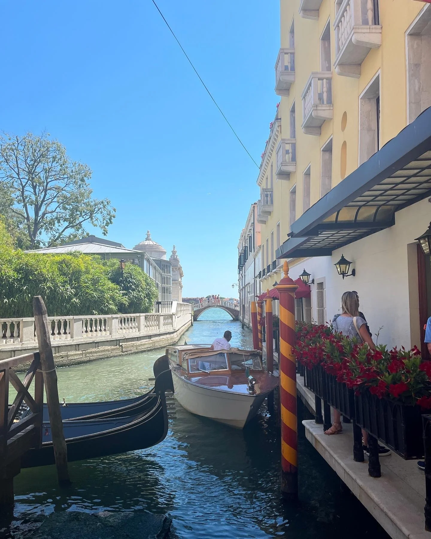 A canal in Venice, Italy, with boats docked along the edge, colorful buildings with balconies, a stone bridge in the distance, people walking near the water, and bright sunny weather.