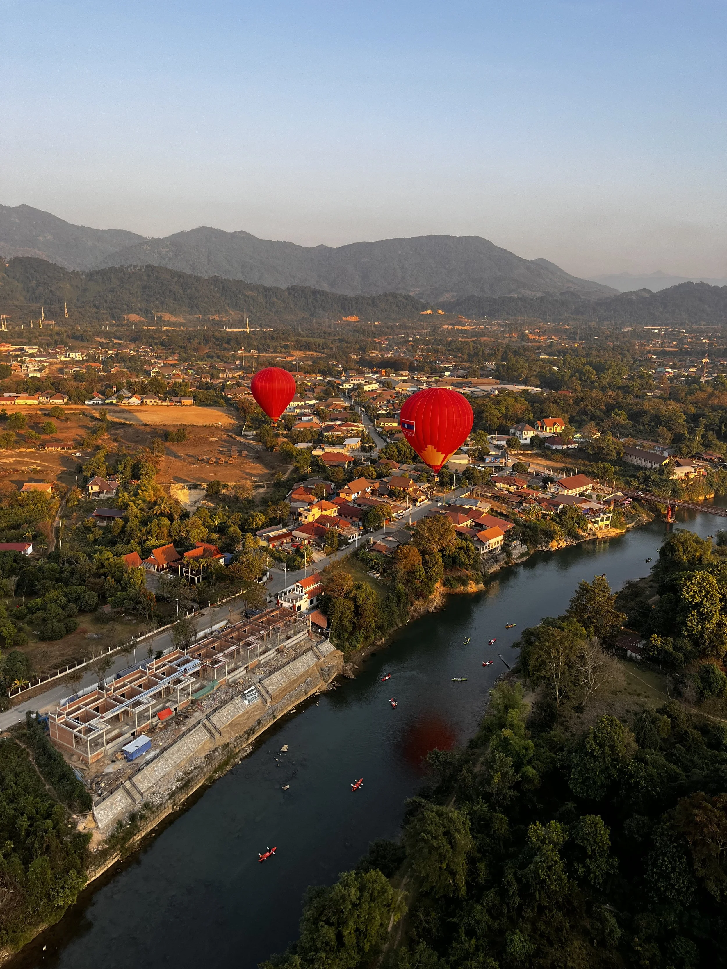 Two red hot air balloons floating over a river with houses and trees along the banks, mountains in the background during sunset.