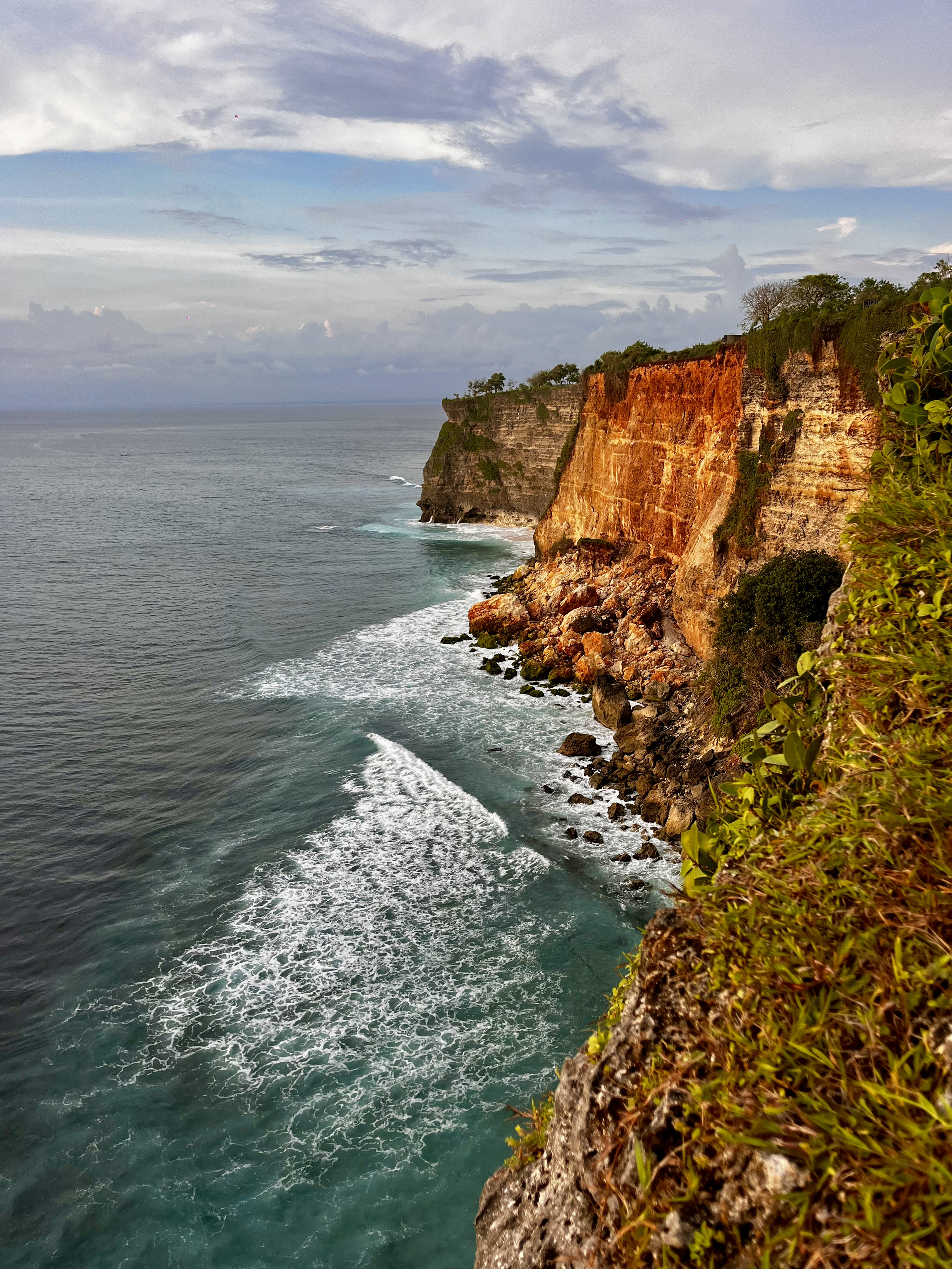 Scenic view of a rugged coastline with high cliffs, green vegetation, and ocean waves crashing against rocks, under a cloudy sky.