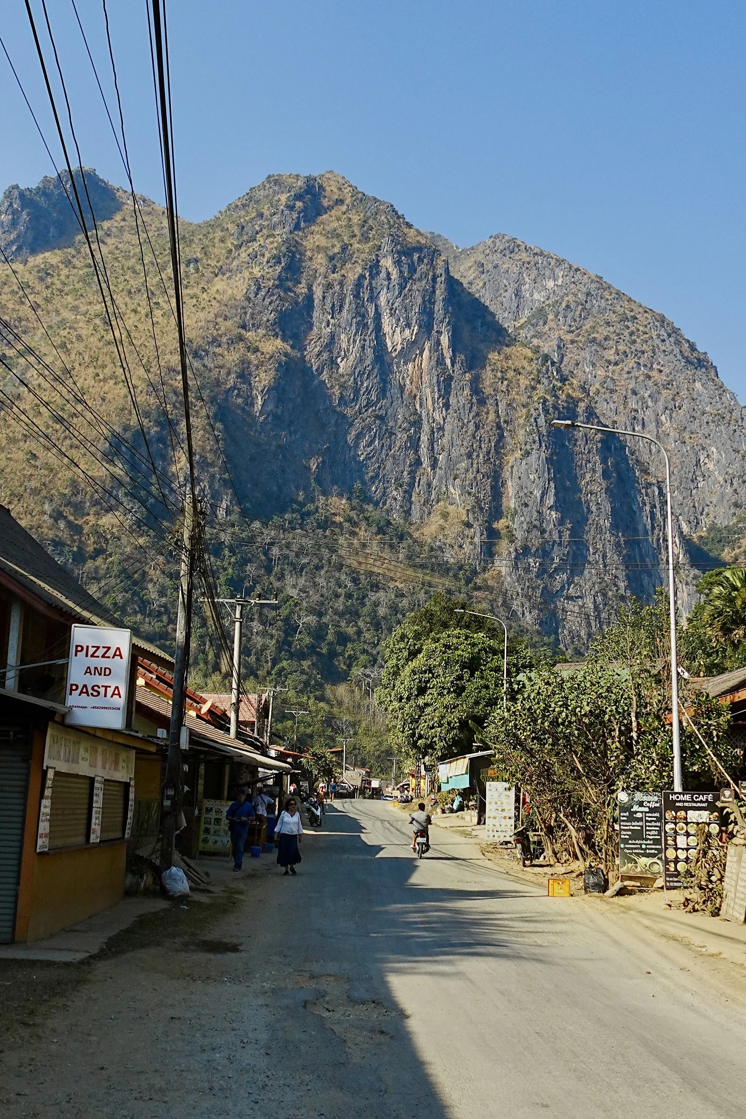 Dirt road in a small town with mountains in the background, a sign for pizza and pasta, and a few people and motorcycles on the street.