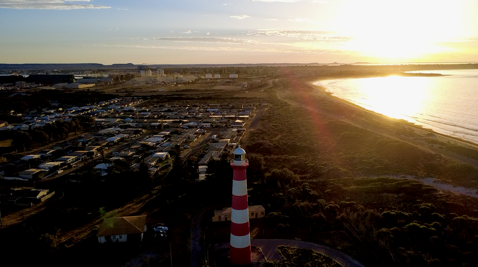 A sunset over a coastal town with a lighthouse with red and white stripes in the foreground, a residential area, and the beach along the shoreline.
